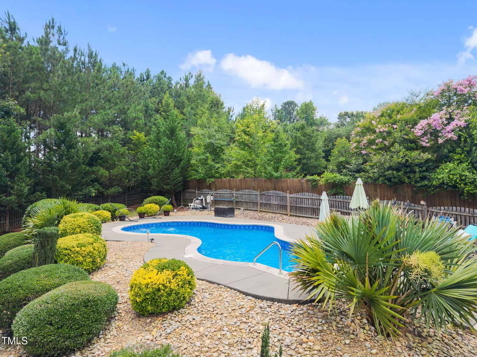 A kidney-shaped swimming pool is surrounded by a landscaped garden with shrubs, small trees, and rocks. A wooden fence and tall trees provide privacy. Two patio umbrellas and lounge chairs are visible near the pool.