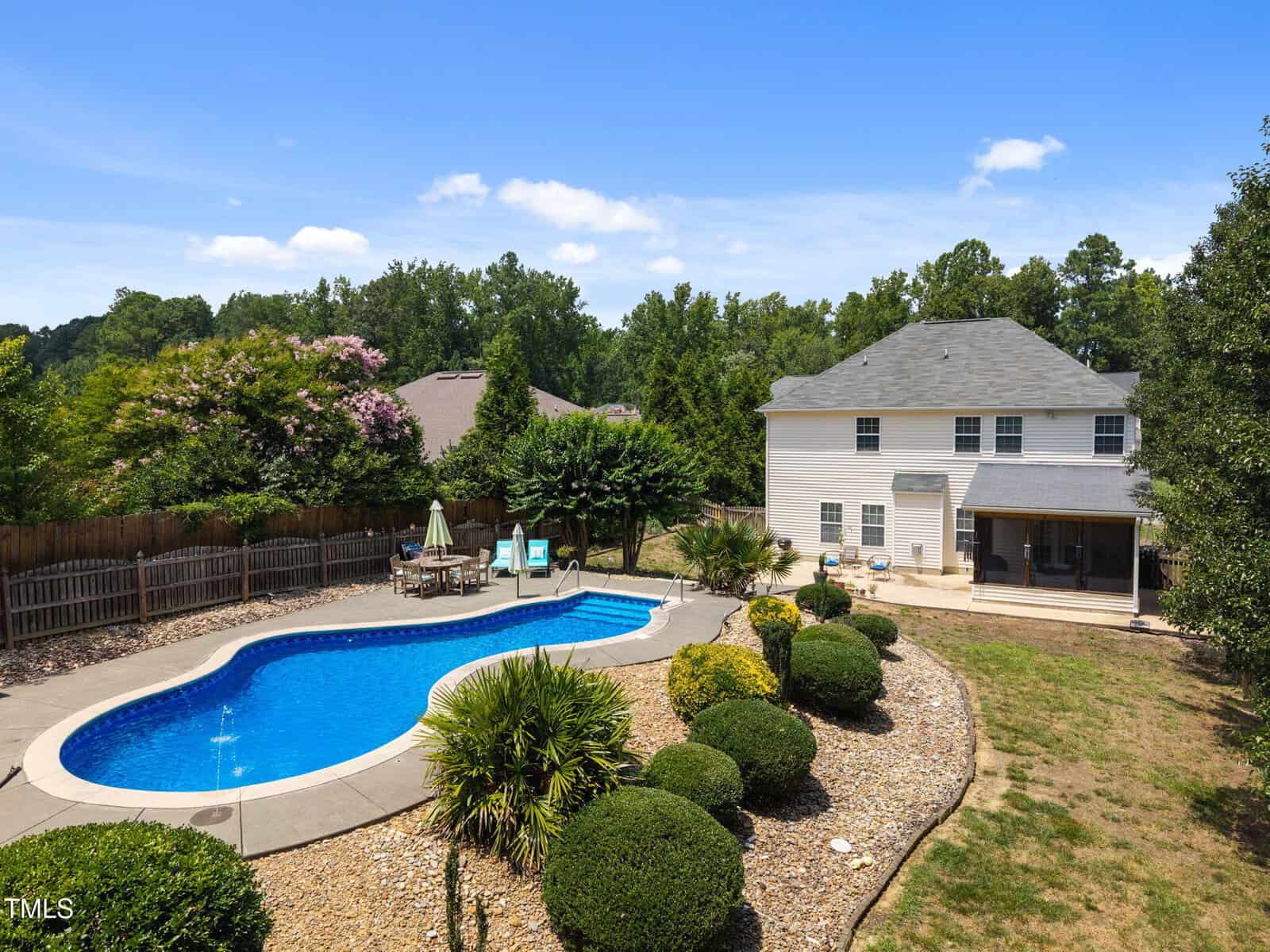 A two-story house with a gray roof and screened patio overlooks a backyard featuring a kidney-shaped in-ground pool, a concrete pool deck, lounge chairs, and landscaped shrubs, with a wooden fence and trees in the background.