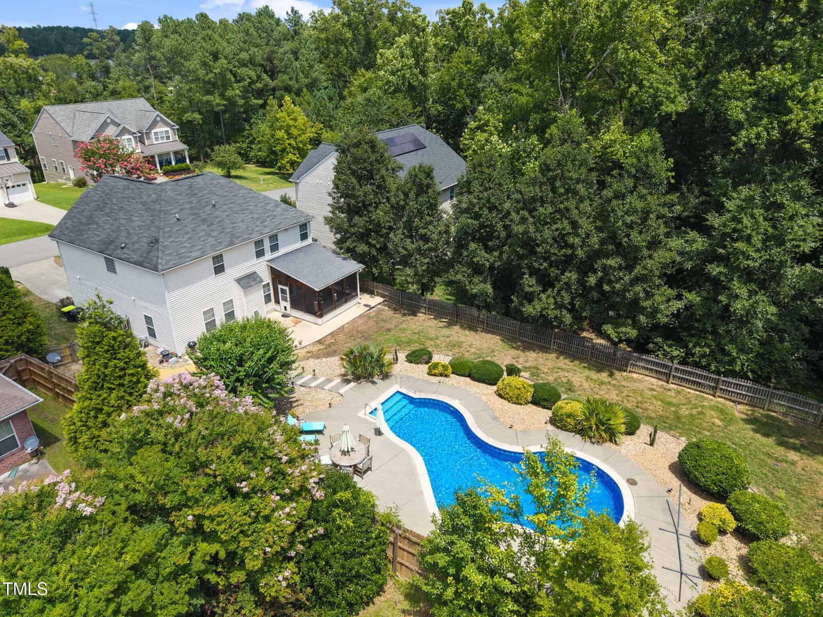 Aerial view of a two-story white house with a fenced backyard featuring a swimming pool, patio area with seating, landscaped bushes, and surrounding trees in a suburban neighborhood.