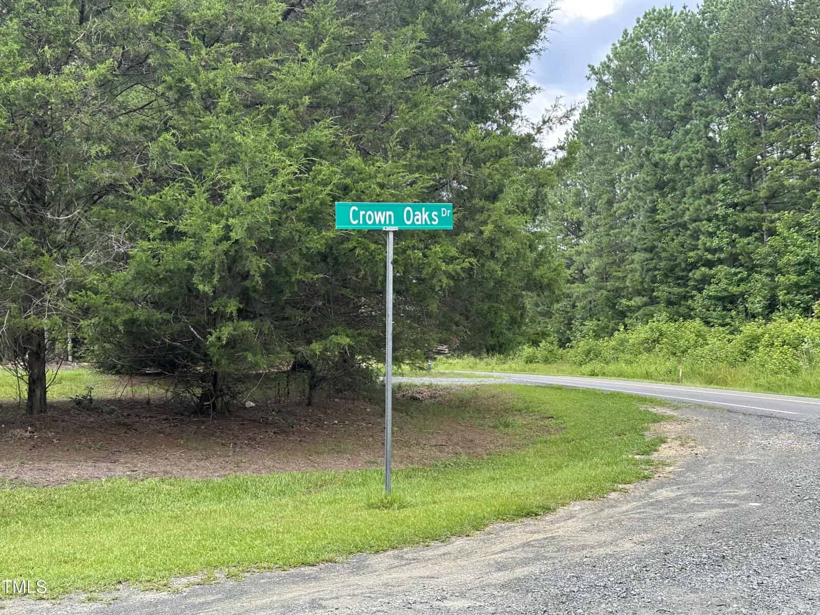 A green street sign labeled "Crown Oaks Dr" stands next to a curved gravel road, with grass and trees in the background on a partly cloudy day.