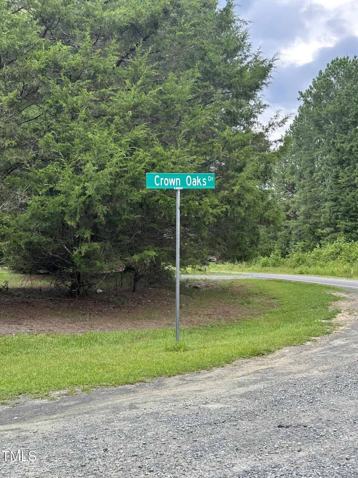 A green street sign reading "Crown Oaks Dr" marks Lot 17 Crown Oaks Drive in Oxford, standing on a grassy area beside a gravel road, with tall green trees and a cloudy sky in the background.