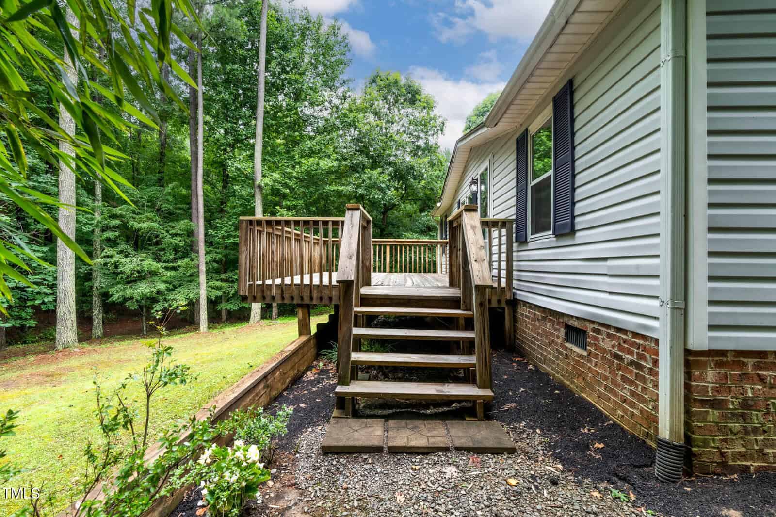 Wooden steps lead up to a small raised deck attached to the side of a house with light siding and brick foundation. The deck overlooks a wooded backyard with green trees and shrubs.