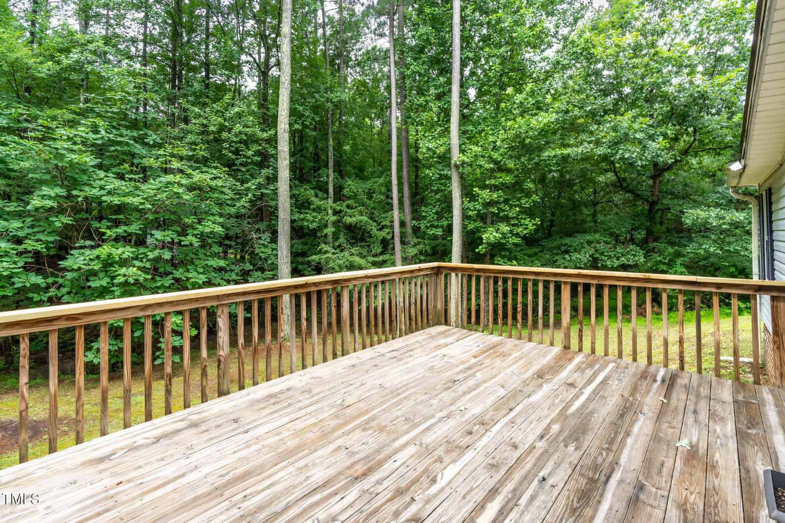 A wooden deck with a simple railing overlooks a yard bordered by tall trees and dense green foliage on a cloudy day.