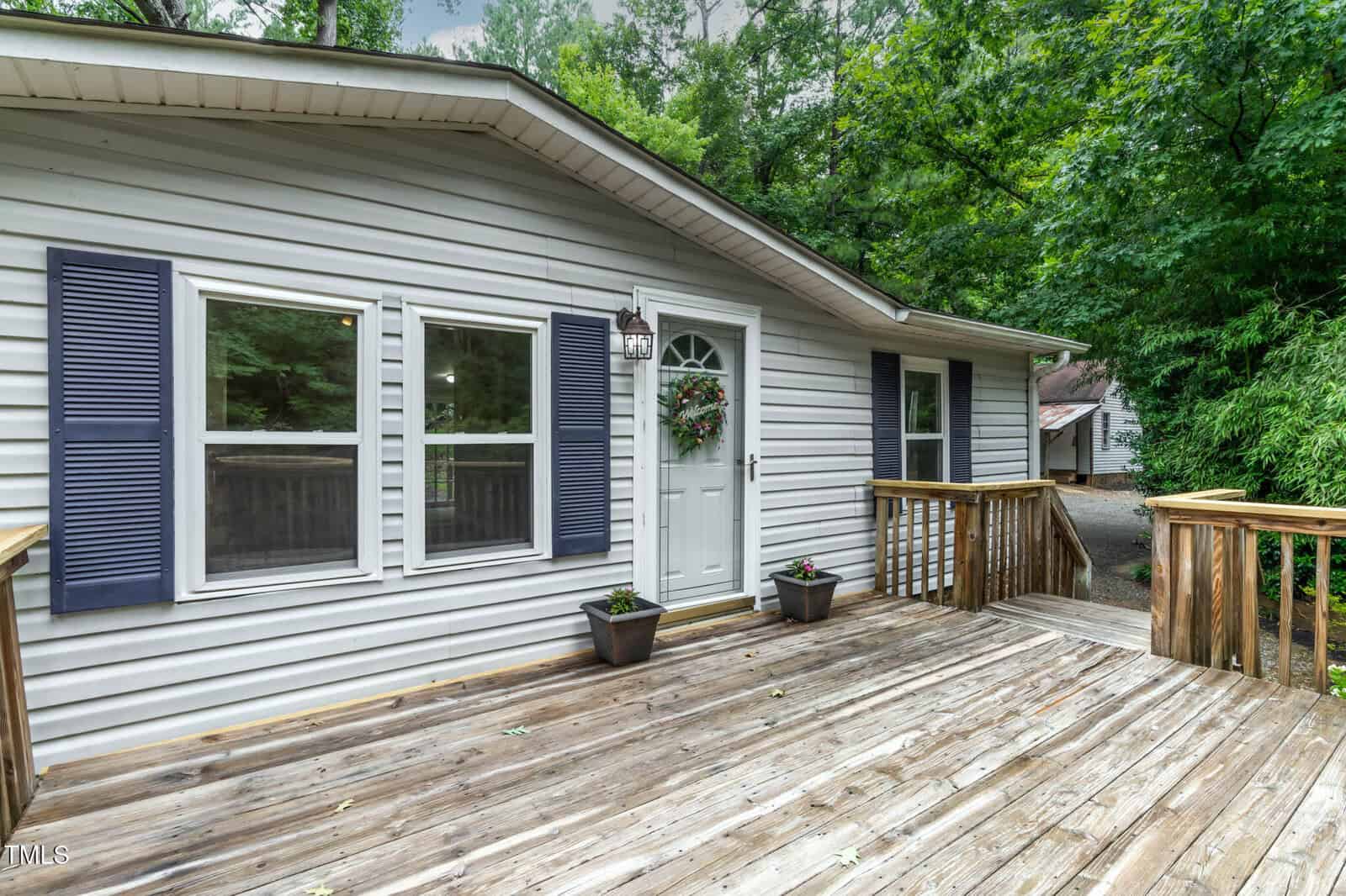 A light gray house with blue shutters, a white door with a wreath, and two potted plants on a wooden deck surrounded by trees. A driveway and another building are visible in the background.