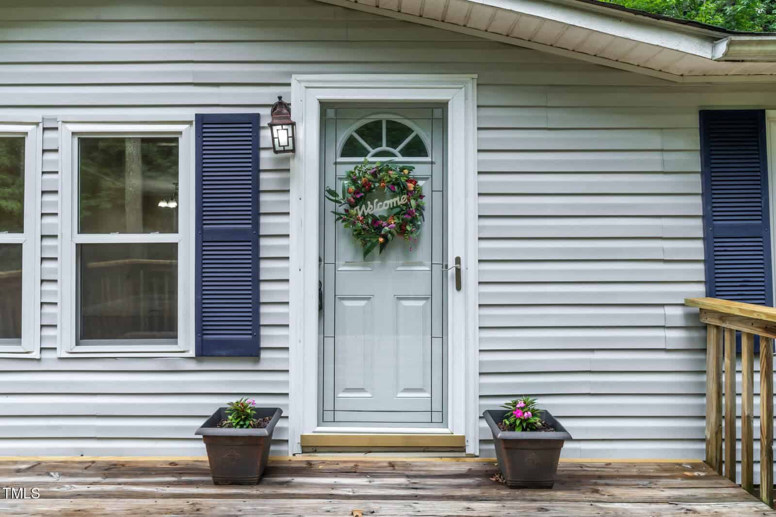 A white house exterior with blue shutters, two windows, and a glass front door. The door has a wreath with the word "Welcome" on it. Two potted plants sit on the wooden porch by the entrance.