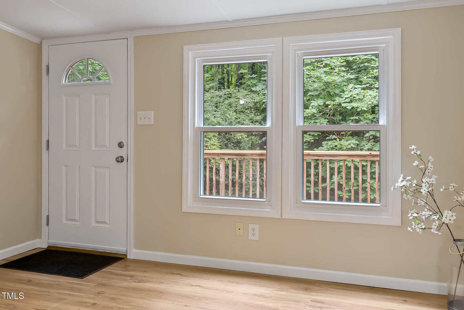 A white entry door with a semi-circular window is next to two double-hung windows overlooking a wooden deck and green trees. The room has beige walls, light wood flooring, and a small vase with white flowers.