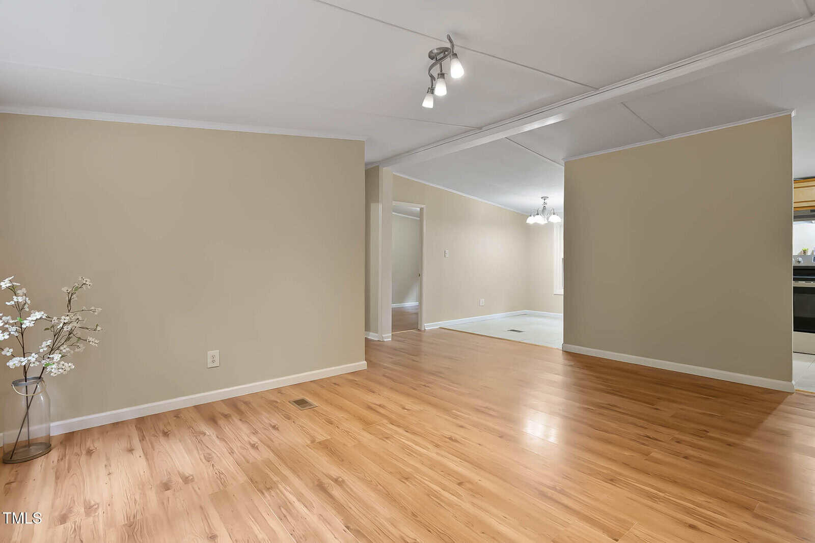 A spacious room with light wood flooring, beige walls, ceiling lights, and a white trim. There is a decorative vase with branches in the corner and an adjoining area with a hanging light fixture visible.