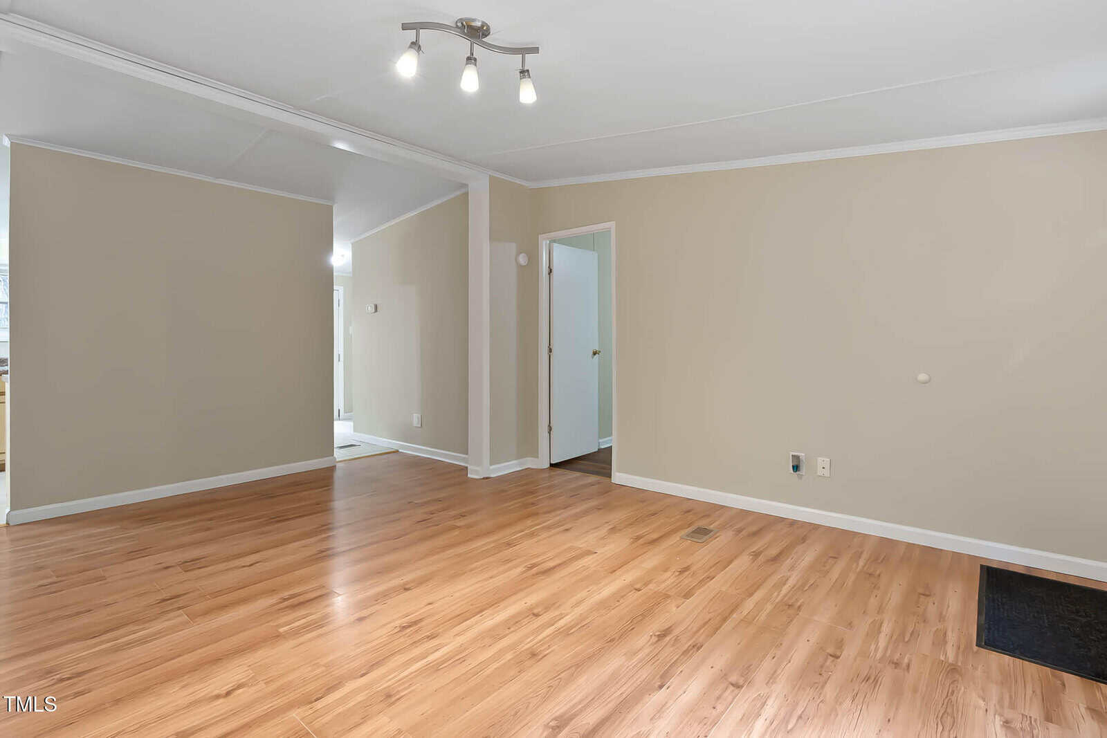 Empty room with light wood flooring, beige walls, white trim, and a modern ceiling light fixture. There are two doorways, one leading to a hallway and the other to another room. Electrical outlets are visible on the wall.