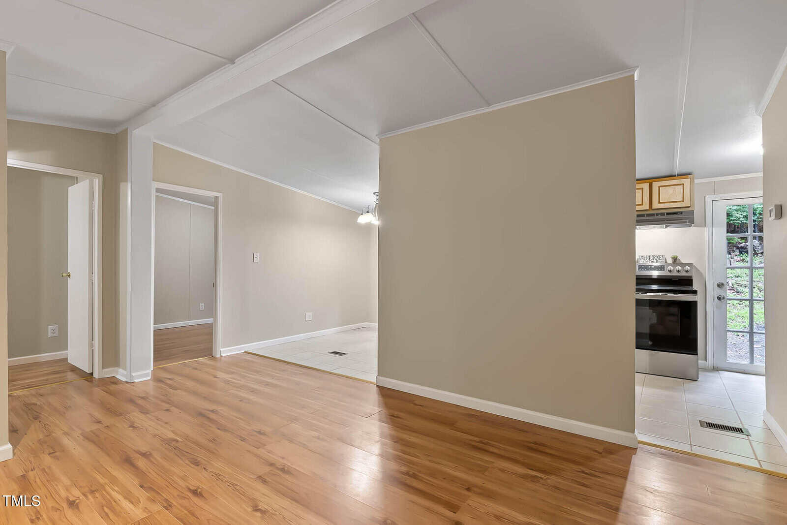 Bright, unfurnished room with light wood floors, beige walls, white trim, an open doorway to a kitchen with tile flooring, and glass doors revealing greenery outside. Ceiling light fixture hangs near the kitchen area.