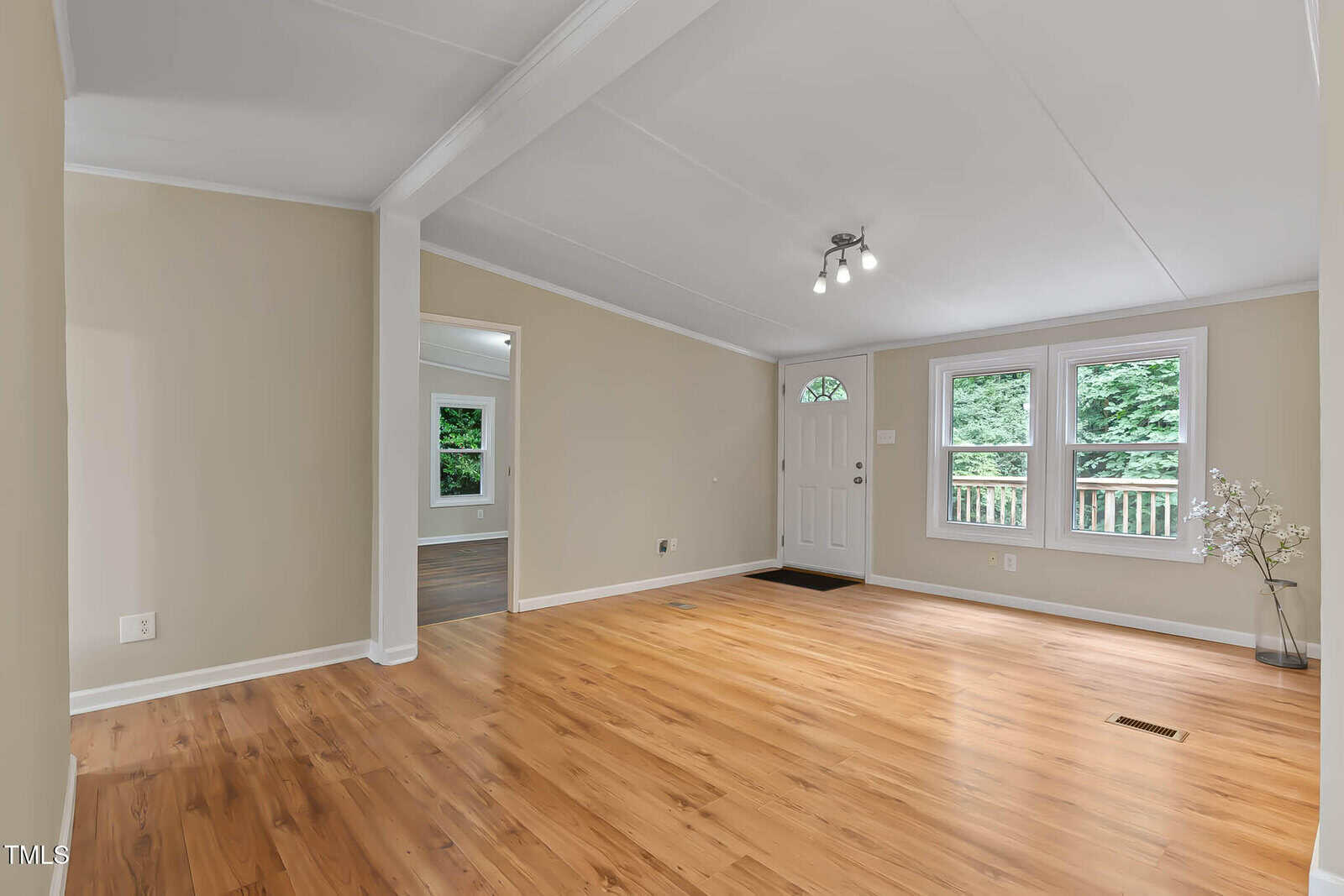 Bright, empty living room with wood floors, beige walls, white ceiling, two windows, front door, and a small potted plant in the corner. Doorway leads to another room with darker wood flooring.
