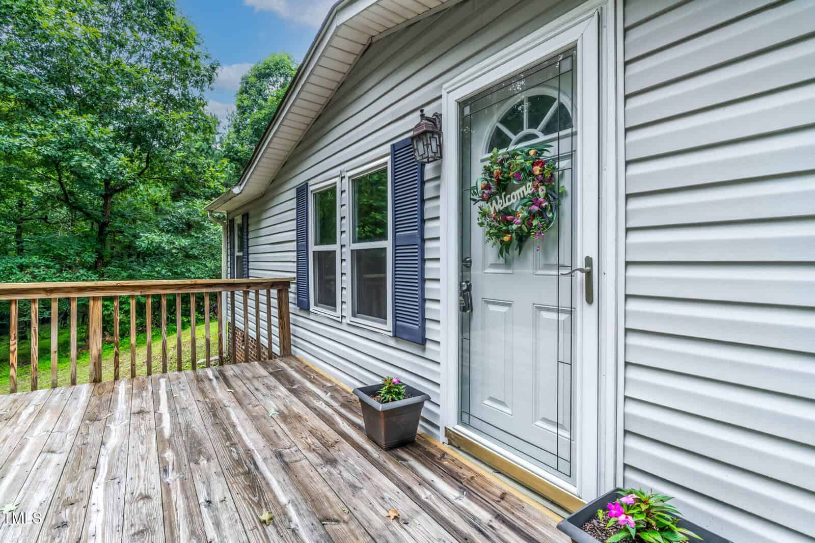 A wooden deck leads to a white front door with a floral wreath and "Welcome" sign. The house has light gray siding, blue shutters, and two potted plants near the door. Lush green trees are visible in the background.