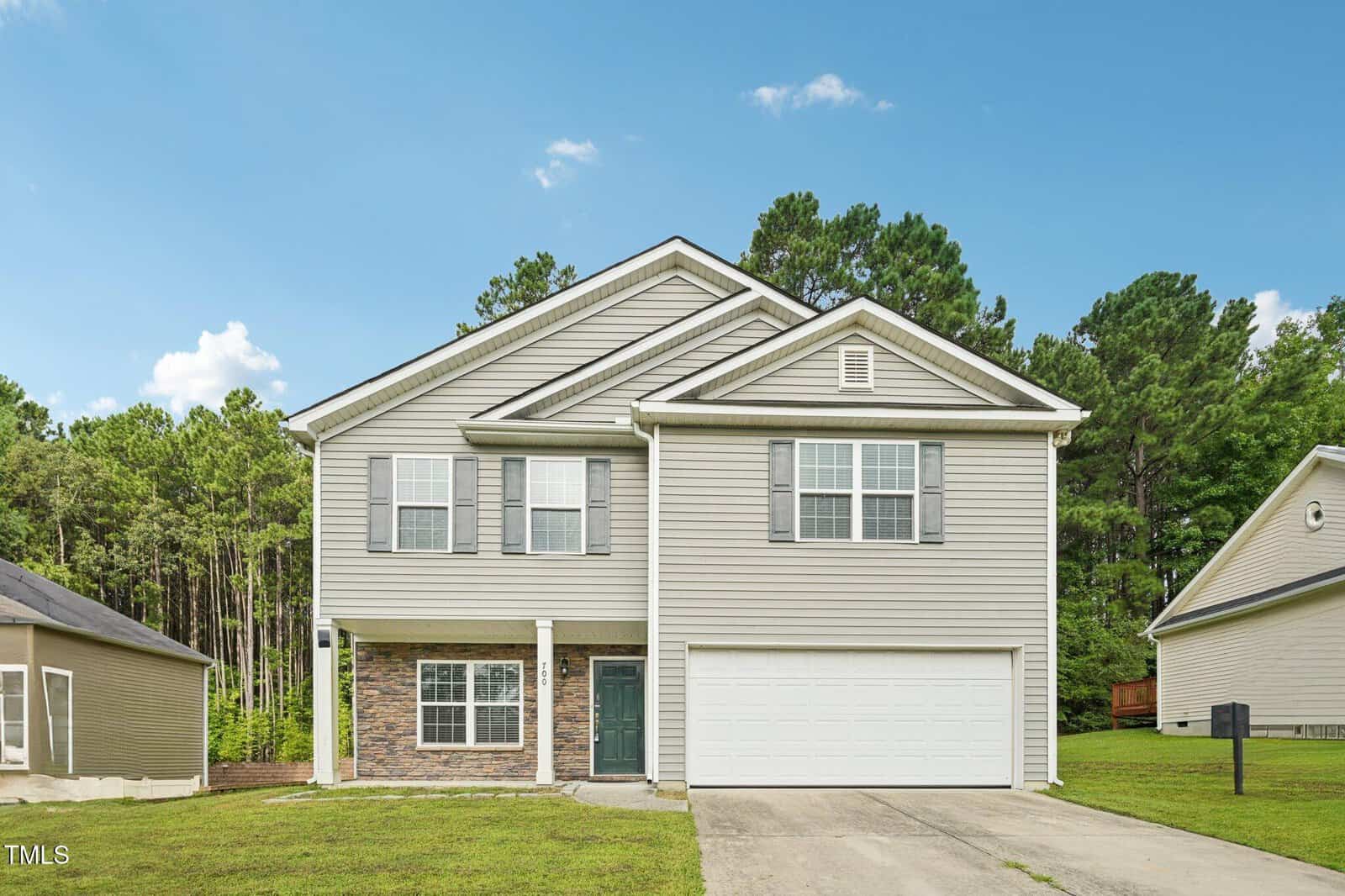 Charming two-story suburban house at 700 Quail Court in Creedmoor, featuring light gray siding, white trim, and a double garage. The front yard has grass and a concrete driveway. Trees and neighboring houses complete the scene.