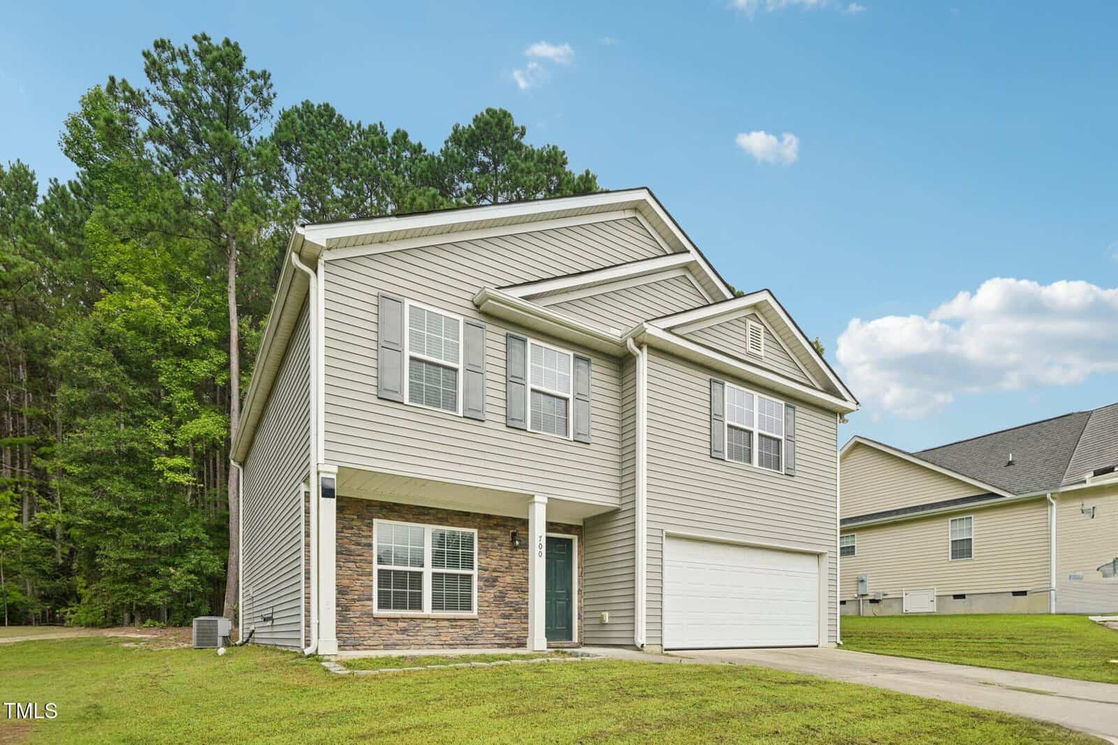 A two-story suburban house with beige siding, white trim, and a double garage. The home has several windows, a small covered entryway, and sits on a grassy lawn with trees in the background.