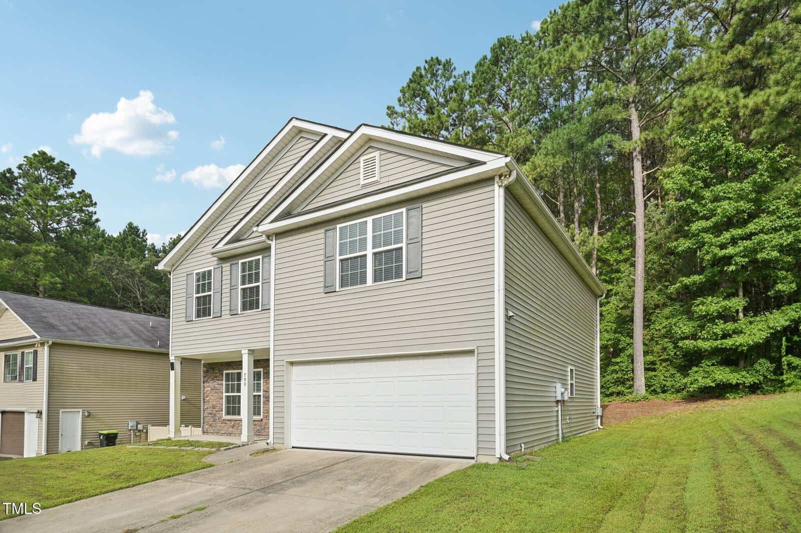 A two-story beige house with white trim and a double garage is shown. The driveway slopes upward, and the house is adjacent to a grassy yard and trees. The sky is blue with a few clouds.
