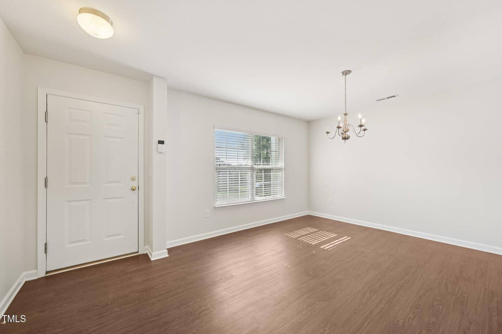 A bright, empty room with light-colored walls, a wood floor, a window with blinds letting in natural light, a simple chandelier, and a white door.
