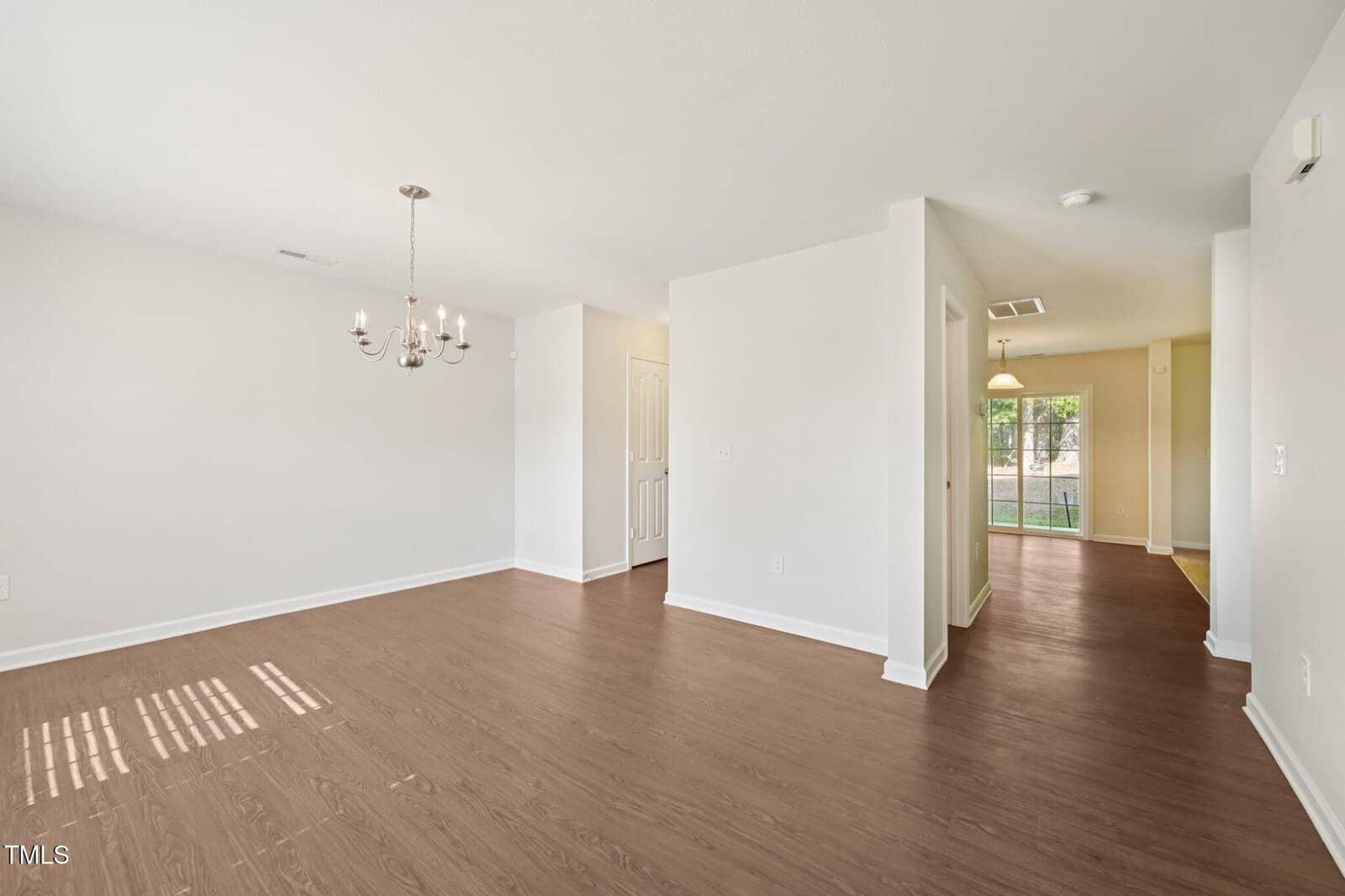A spacious empty room with light-colored walls, a wood floor, a modern chandelier, and natural light coming from windows. A hallway leads to another area with a glass door opening to the outside.
