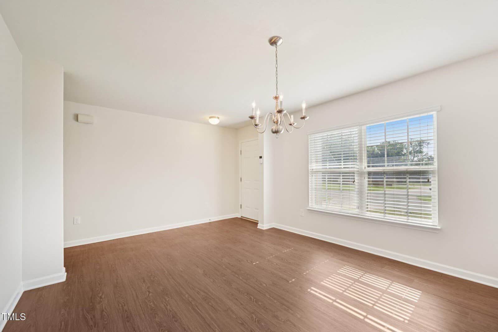 An empty room with light-colored walls, wood flooring, a large window with blinds letting in natural light, a white entry door, and a modern chandelier hanging from the ceiling.
