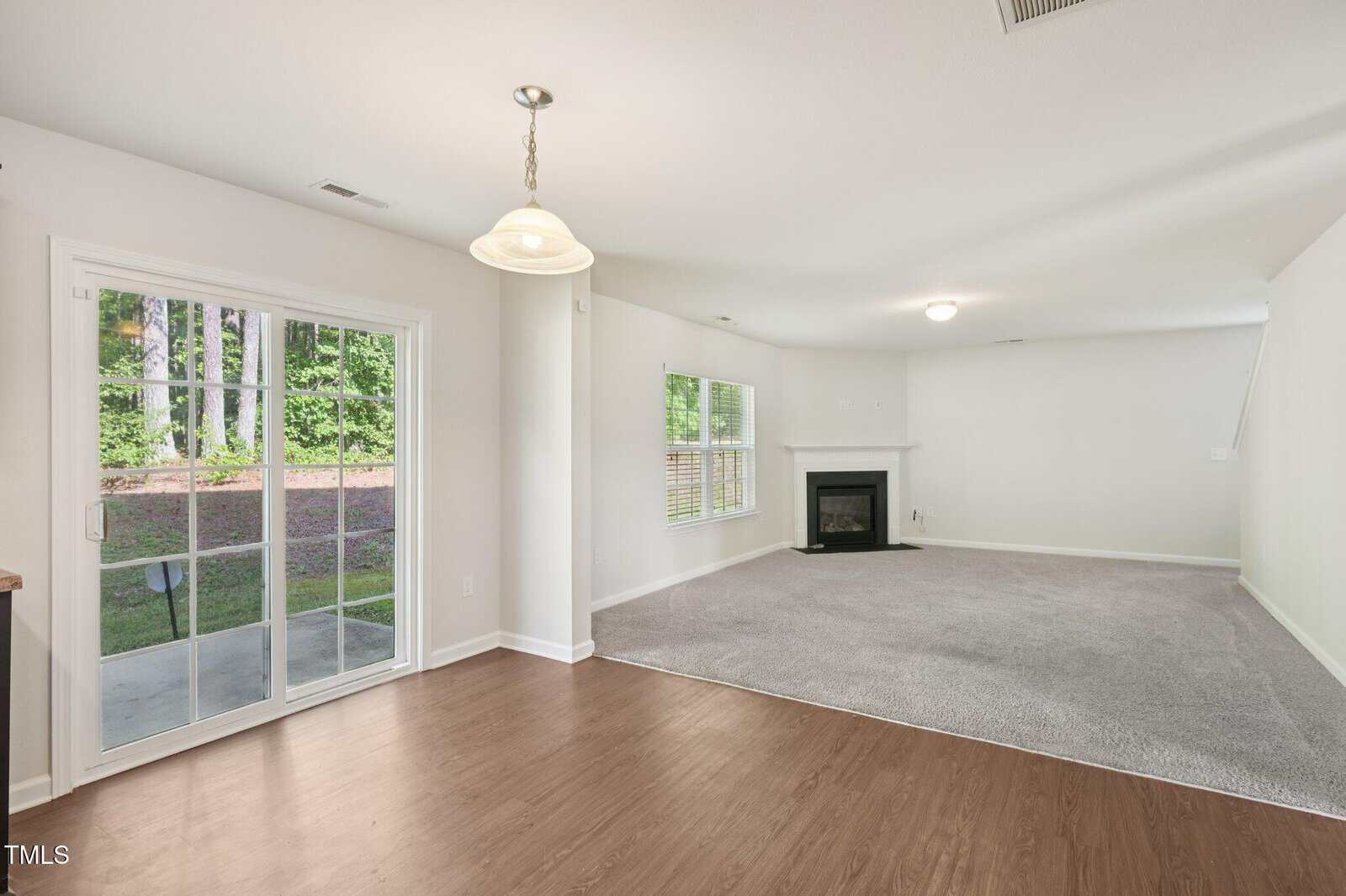 A bright living area with light walls, carpeted flooring, and a corner fireplace, adjacent to a dining area with wood flooring and a hanging light fixture. Sliding glass doors open to a view of trees outside.