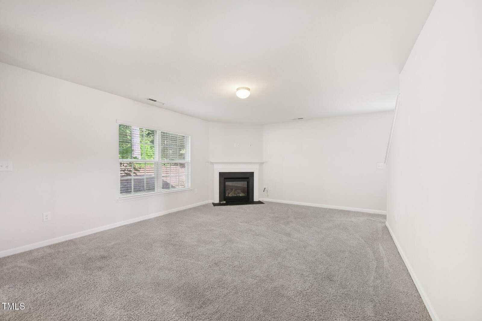A spacious, empty living room with light gray carpet, white walls, a window with blinds, and a corner fireplace. There is a staircase visible on the right side of the image.