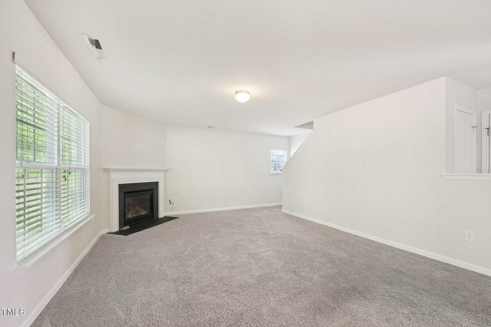 A spacious living room with light gray carpet, white walls, a fireplace on the left wall, and a large window providing natural light. A stairway and a partial view of another room are visible in the background.