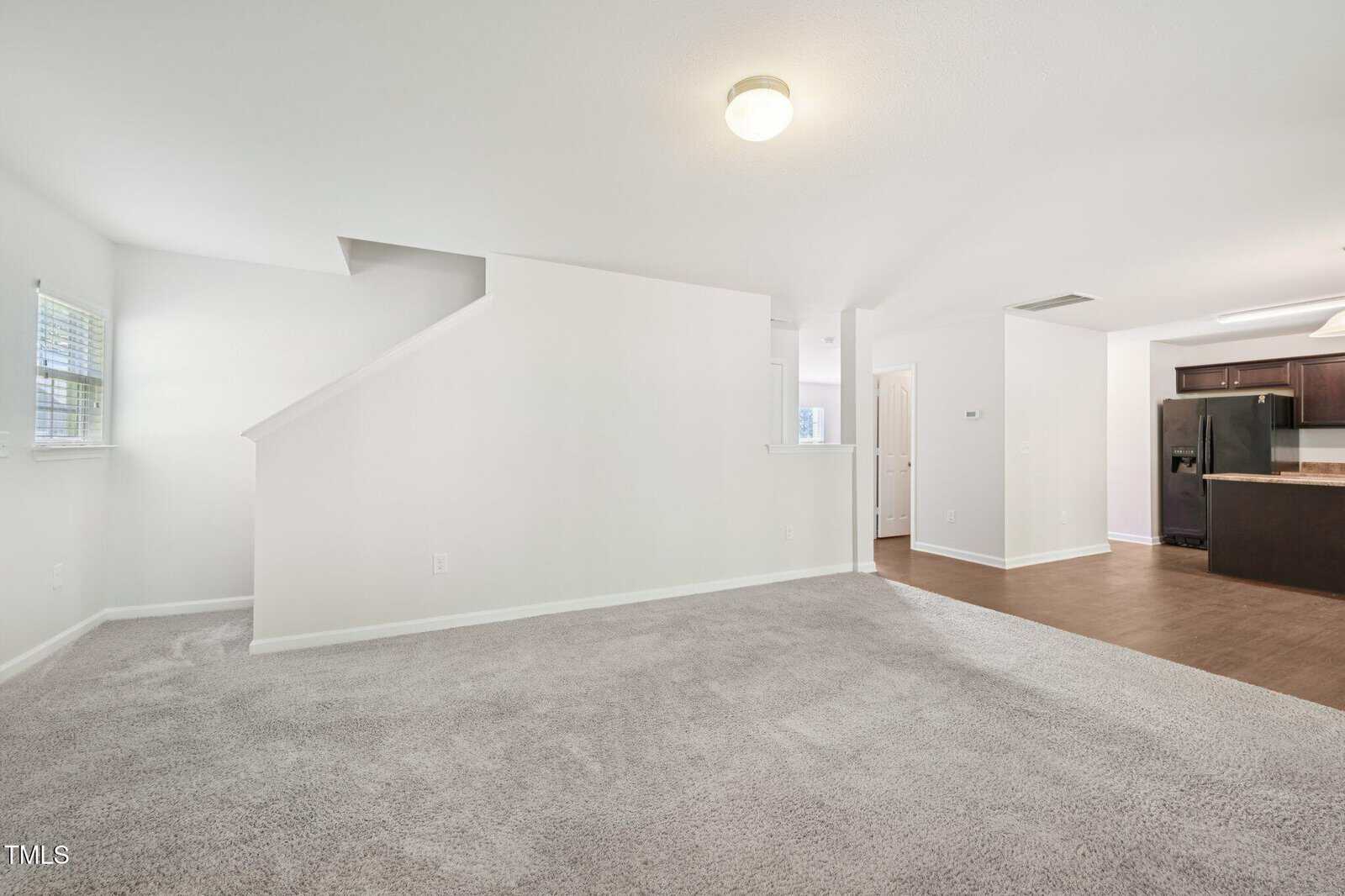 Unfurnished living space with light gray carpet, white walls, and a central staircase. Adjacent area has dark wood flooring, dark kitchen cabinets, and stainless steel appliances. Ceiling light fixture is visible.