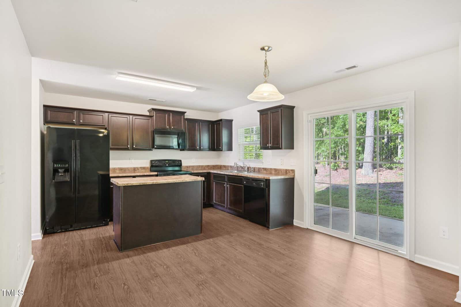 A modern kitchen with dark cabinets, black appliances, brown countertops, wood flooring, a hanging light, and sliding glass doors leading to a patio with a wooded view.