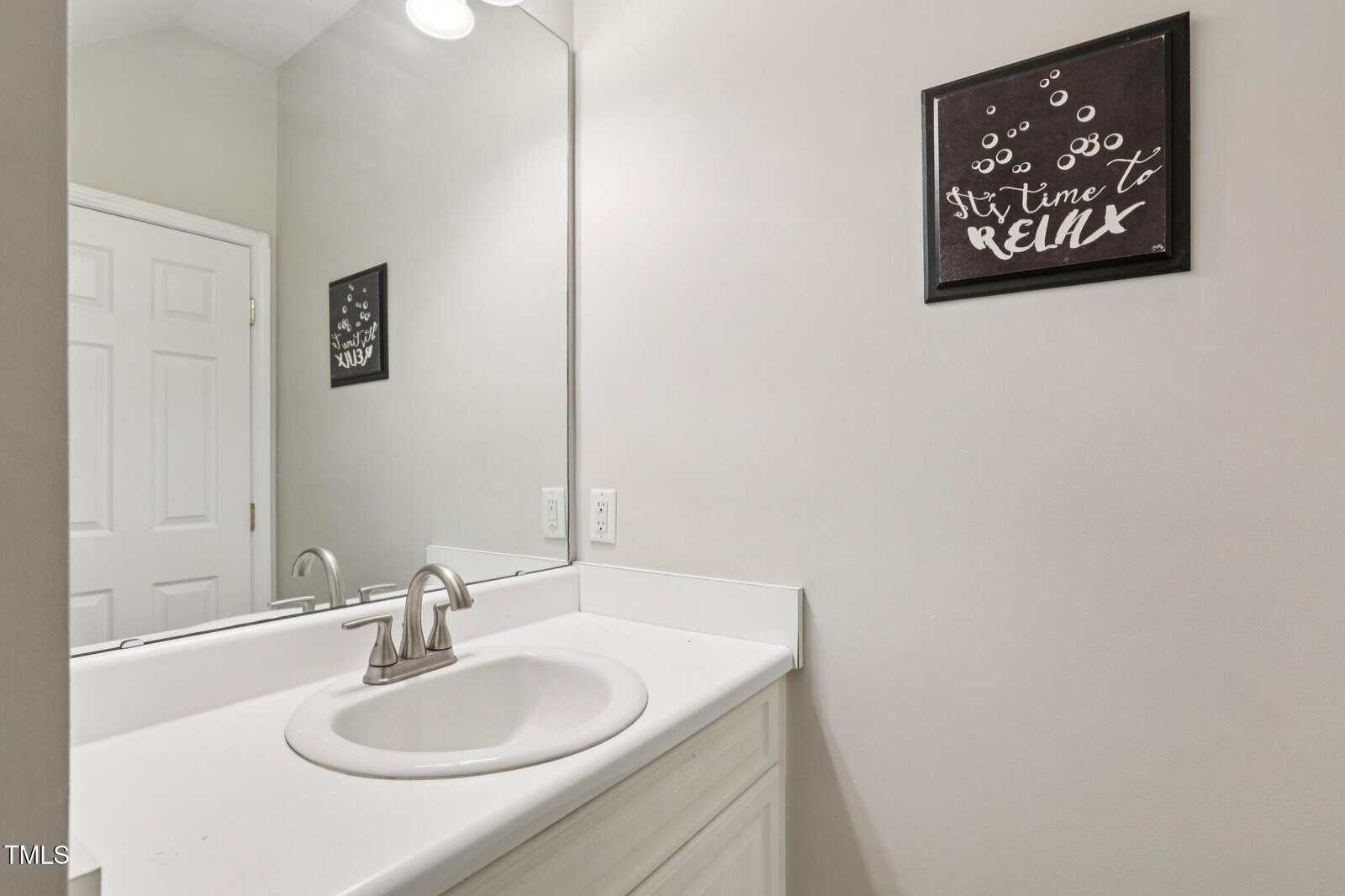 A bathroom with a white sink and countertop, silver faucet, large mirror, and a wall sign that says “It’s time to relax” with bubble designs.