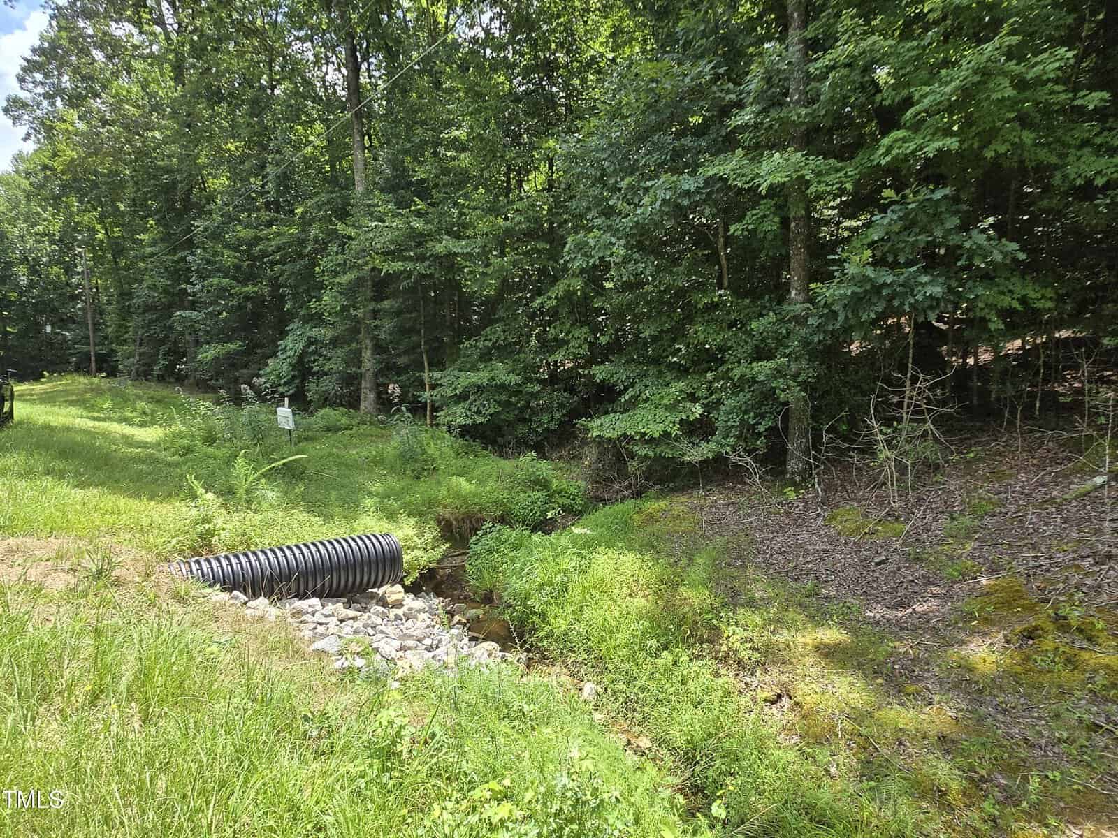 A black corrugated drainage pipe channels water through a grassy ditch beside a forested area, with dense green trees and sunlight filtering through the leaves. Rocks line the streambed around the pipe.