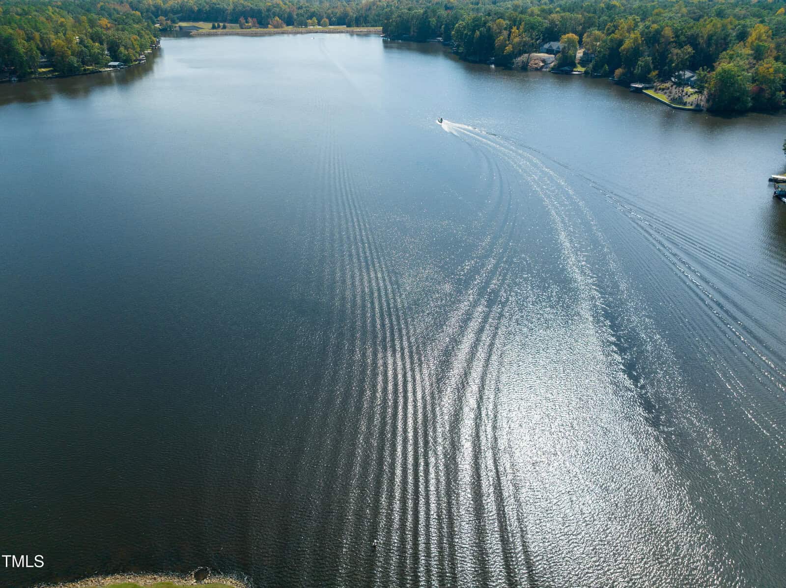 Aerial view of a calm lake with a boat creating rippling waves, surrounded by trees with green and autumn foliage along the shoreline under clear skies.