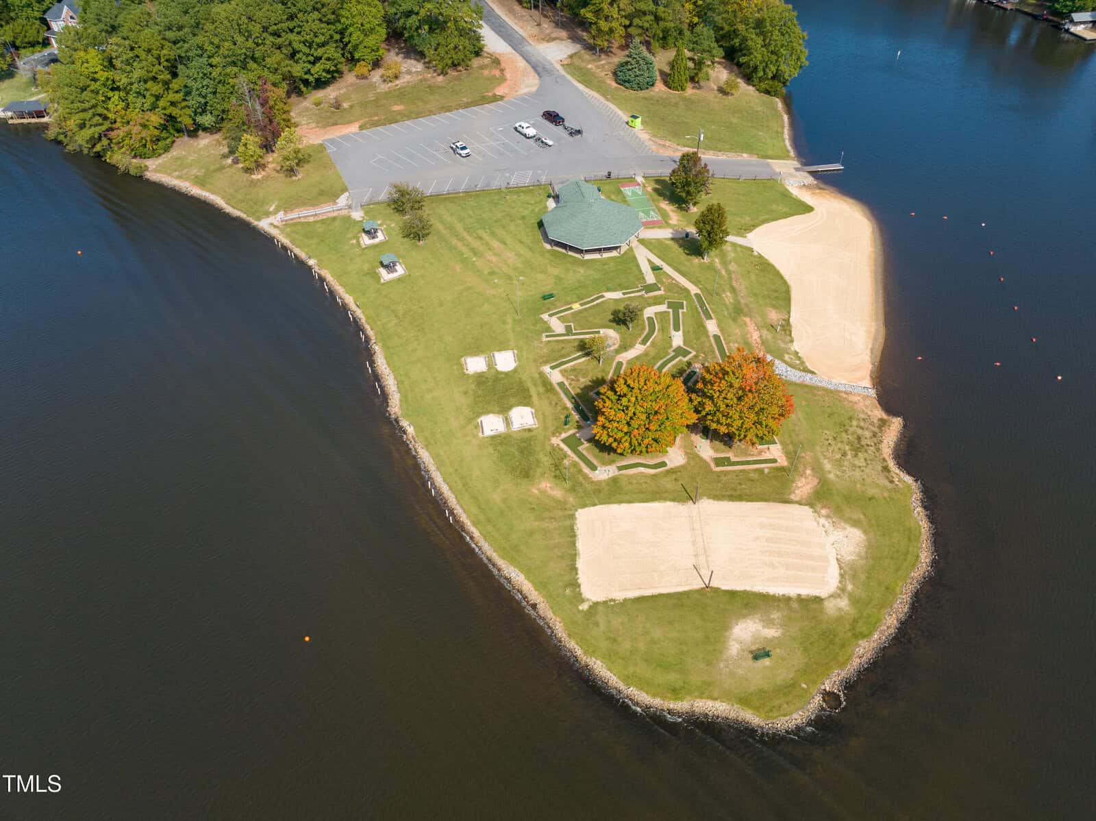 Aerial view of a small park on a peninsula with a gazebo, walking paths, picnic tables, a sandy volleyball court, beach areas, and a parking lot surrounded by water and trees.