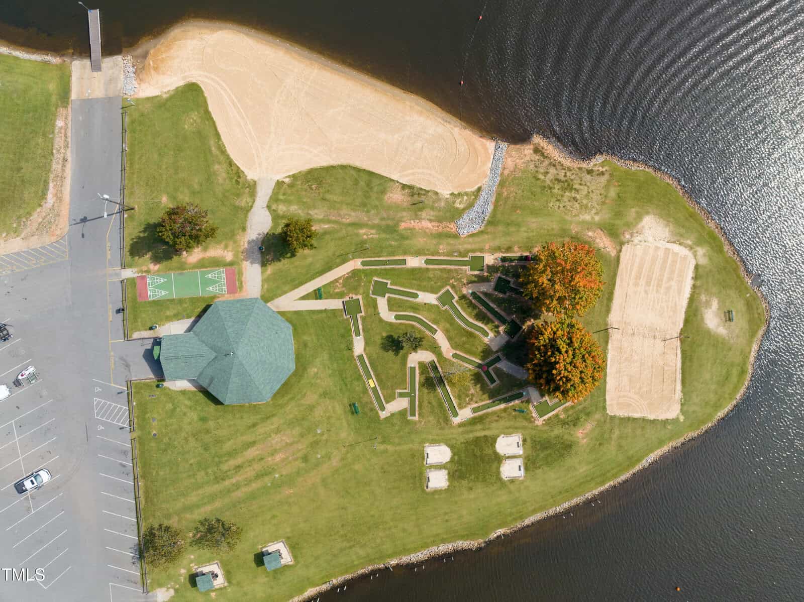 Aerial view of a park by a body of water featuring a pavilion, walking paths, playgrounds, parking lot, sand courts, picnic tables, green lawns, and trees with some autumn foliage.
