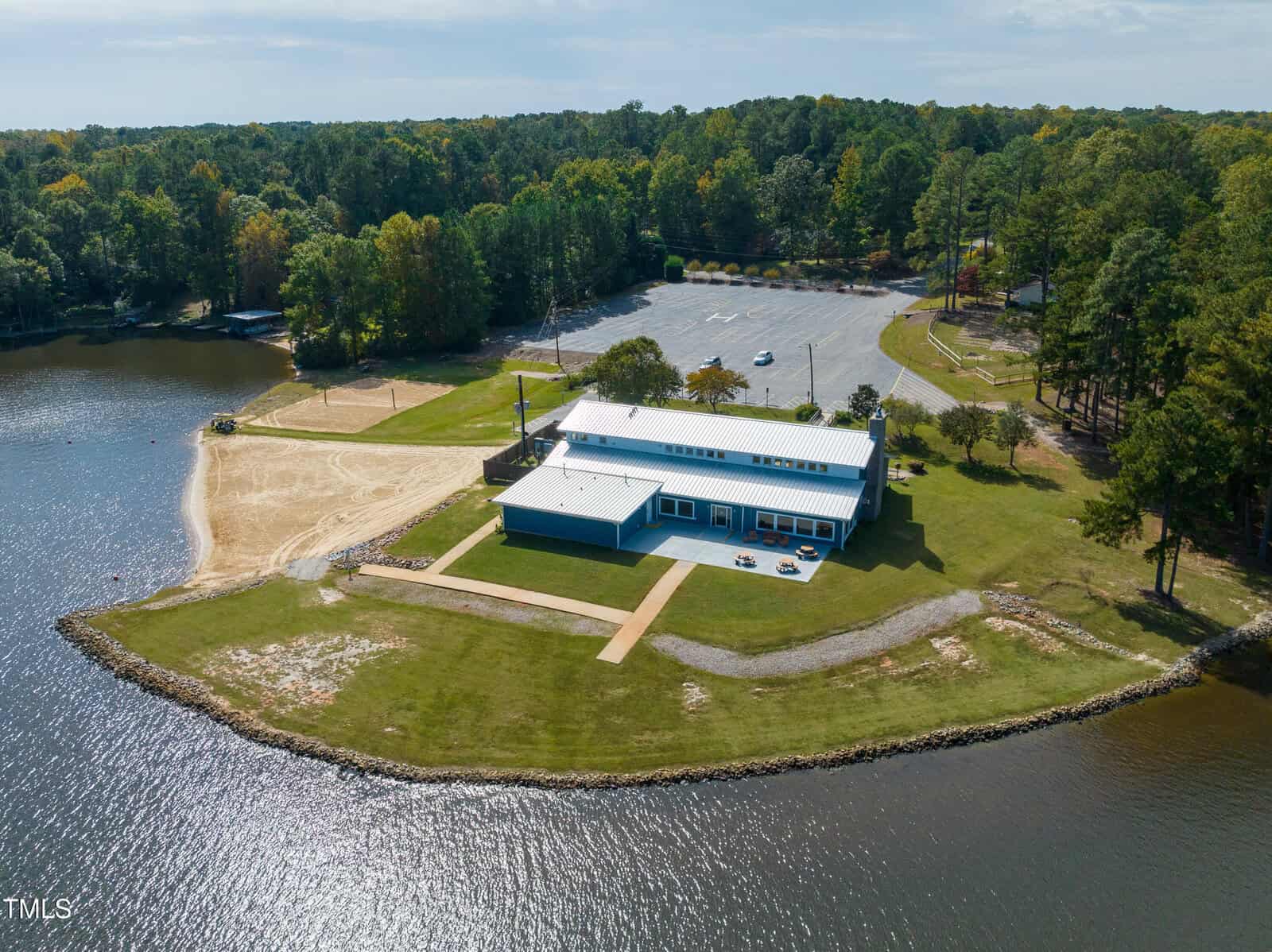 A large building with a white roof sits on a grassy peninsula by a lake, with a sandy beach area, nearby trees, and a parking lot in the background under a partly cloudy sky.