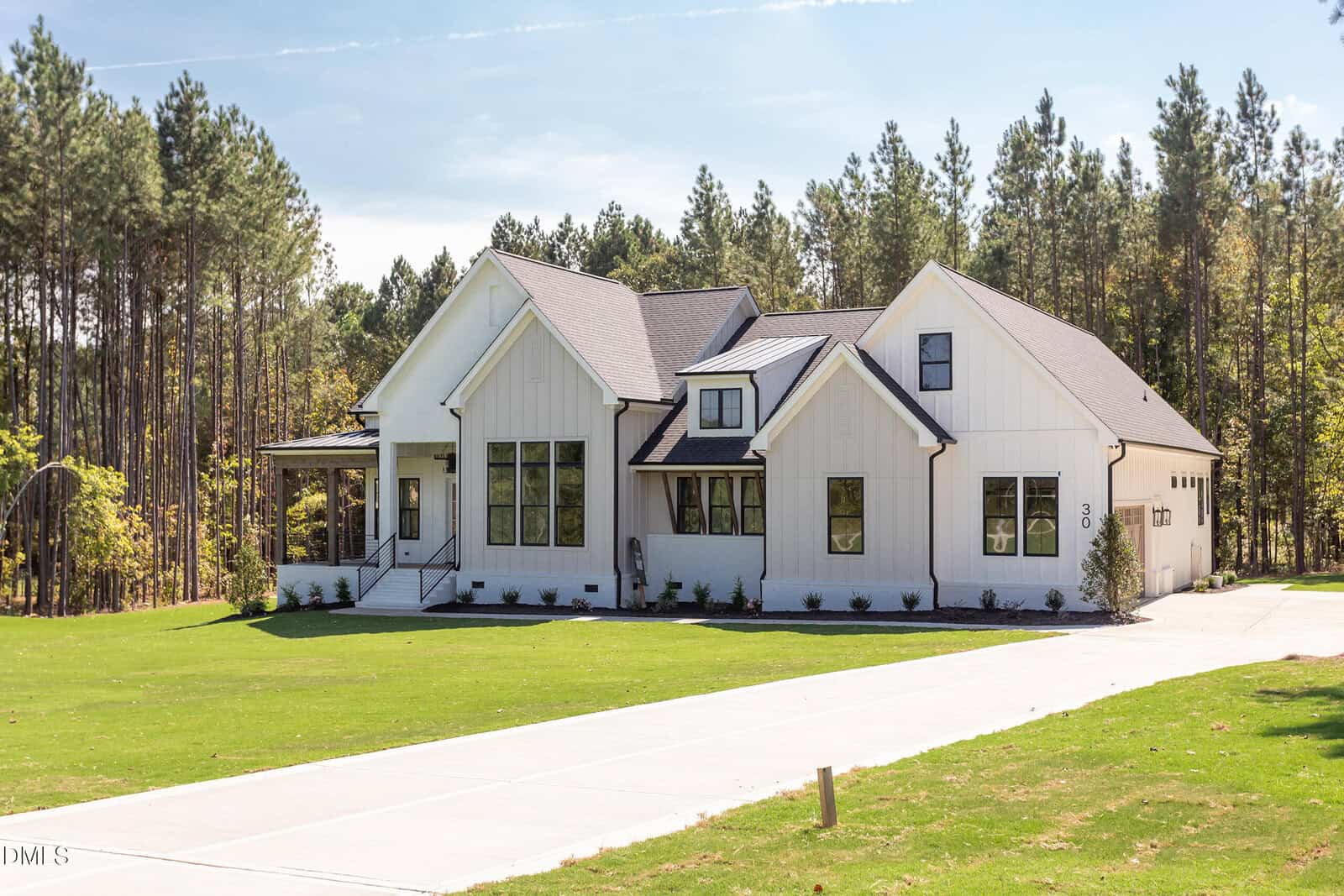 A modern white house with black trim sits on a large, well-manicured lawn, surrounded by tall pine trees. The driveway leads to a three-car garage on the right side of the house. The sky is clear and sunny.