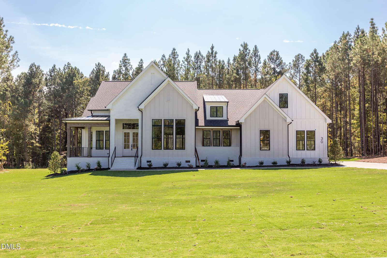 A modern white farmhouse with black-framed windows sits on a large green lawn at 30 Harvest View Way in Franklinton, surrounded by tall pine trees under a clear sky. The house has a front porch, gabled rooflines, and a paved driveway on the right side.