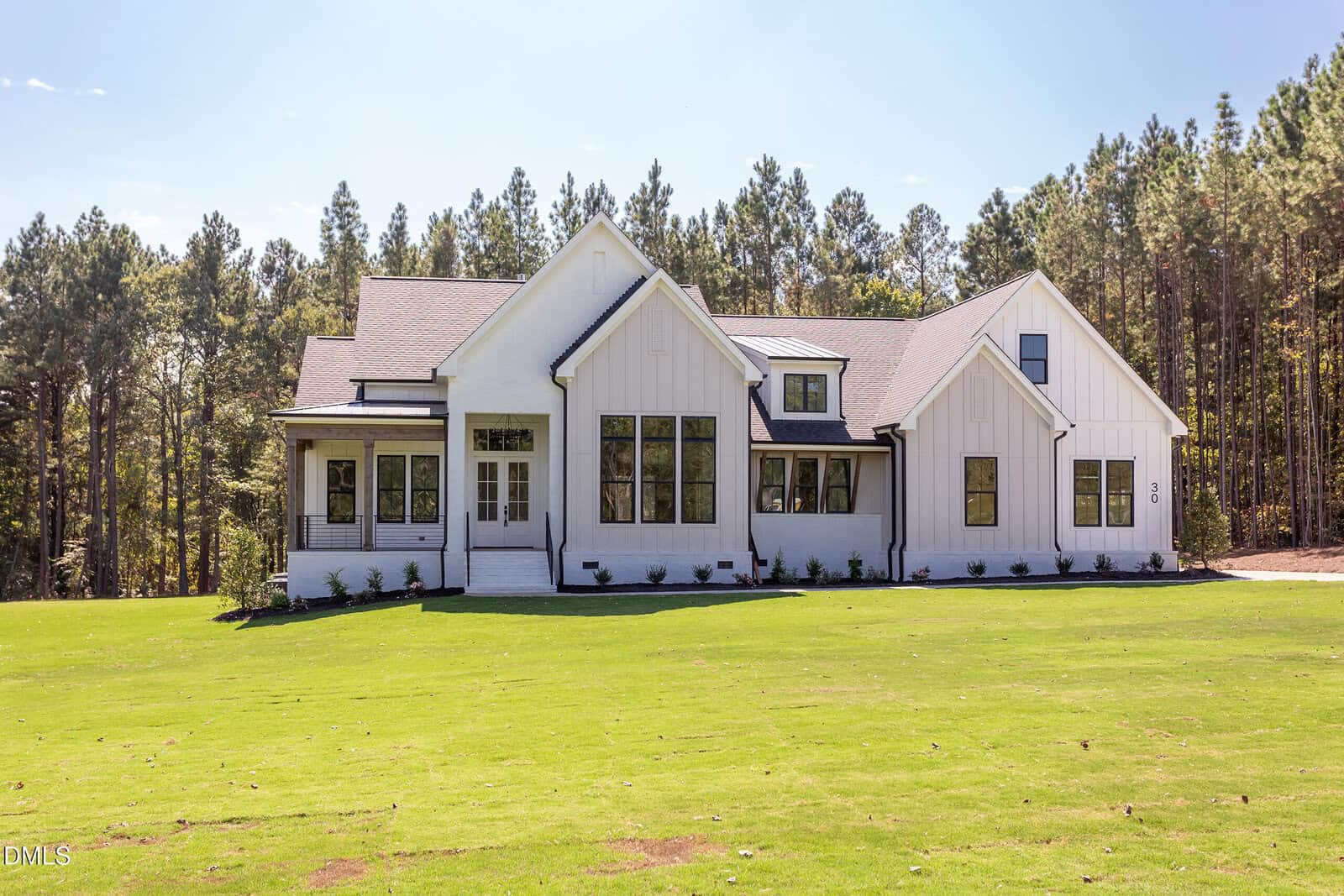 A white modern farmhouse with large windows and a gray roof sits on a grassy lawn, surrounded by tall pine trees under a clear blue sky.