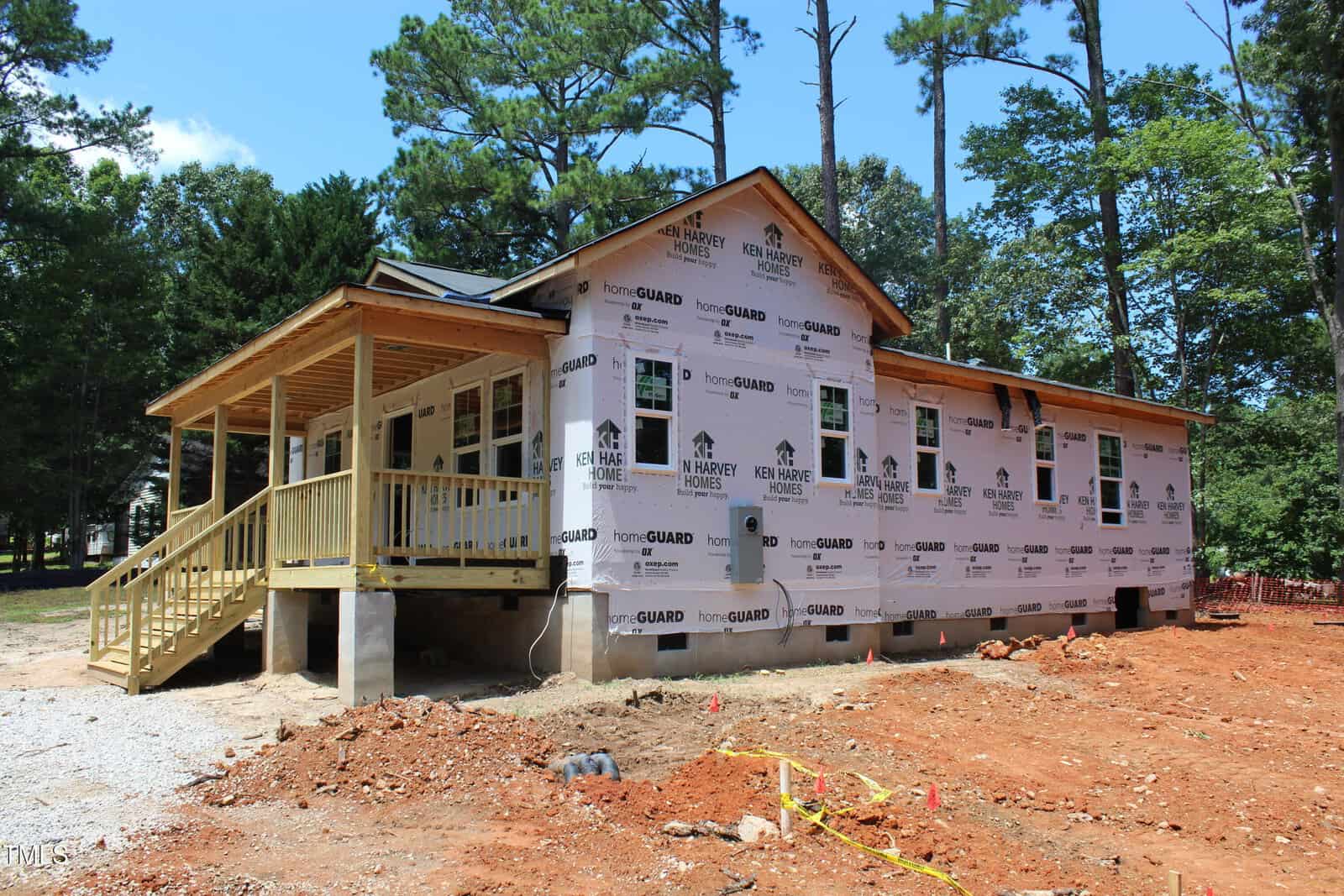 A single-story house under construction, covered in HomeGuard wrap with exposed wooden framing. The ground is bare dirt, and the porch is partially built. Trees surround the site under a clear sky.