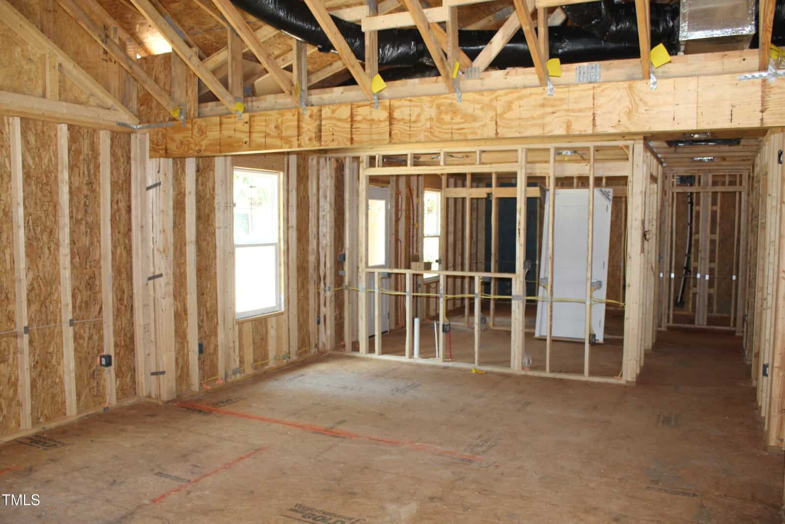Interior view of a house under construction, showing exposed wooden framing, open ceiling beams, insulation ducts, and unfinished floors. There are windows and partial framing for future rooms. No drywall or paint is applied yet.