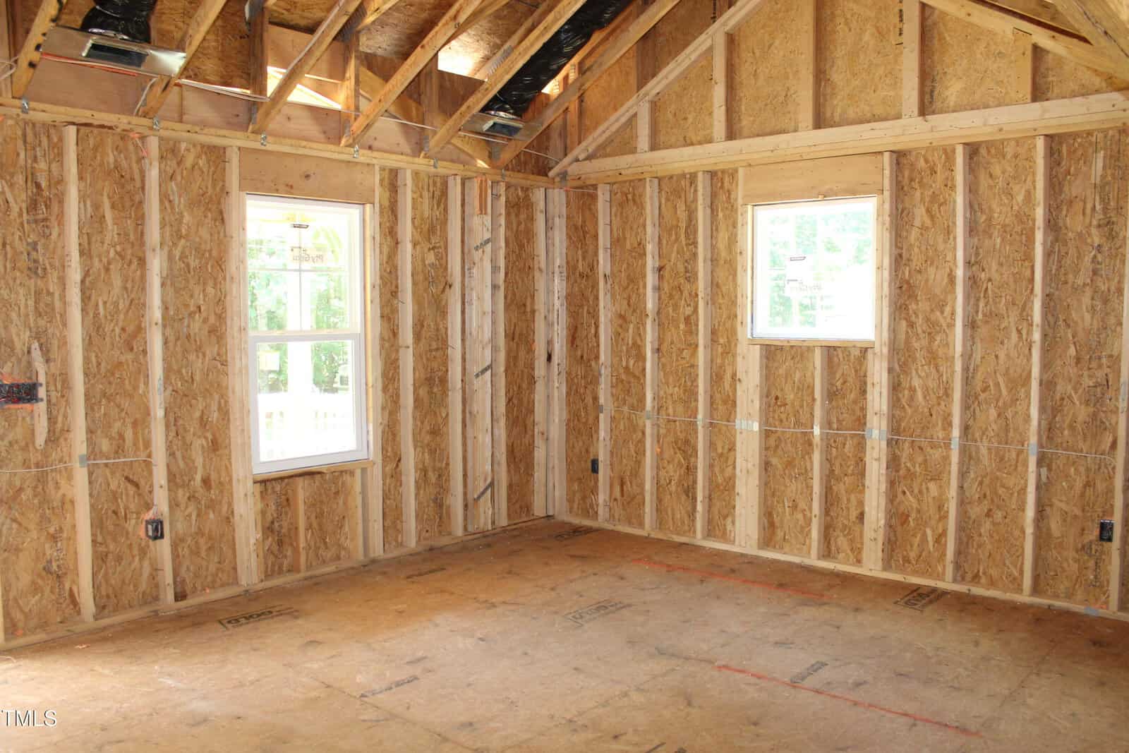 Unfinished room with exposed plywood walls, open ceiling rafters, two windows, and bare subfloor. Sunlight is coming in through the windows, illuminating the construction space.