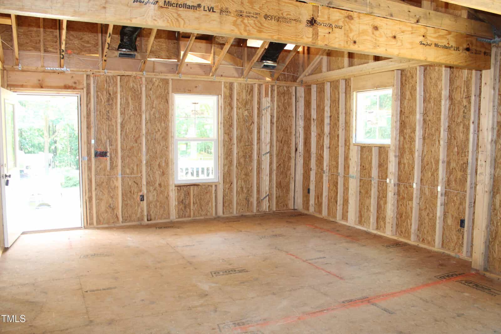 A room under construction with exposed wooden framing, plywood floors, open ceiling beams, two windows, and a door open to the outside. Sunlight comes through the windows and door, illuminating the unfinished space.
