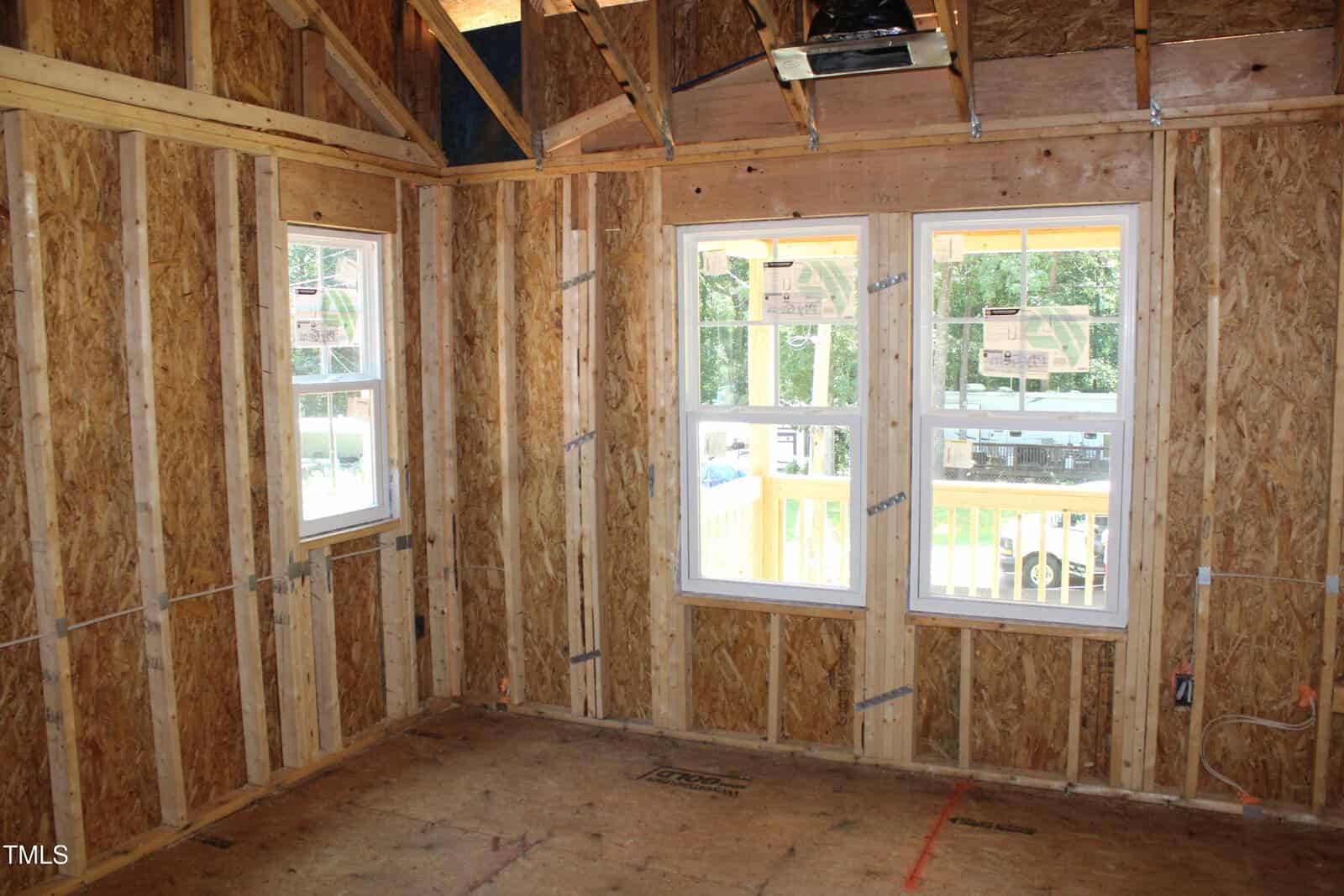 Partially constructed room with exposed wooden framing and insulation, featuring three rectangular windows. Sunlight enters through the windows, illuminating the unfinished interior. Ceiling rafters and electrical wiring are visible.