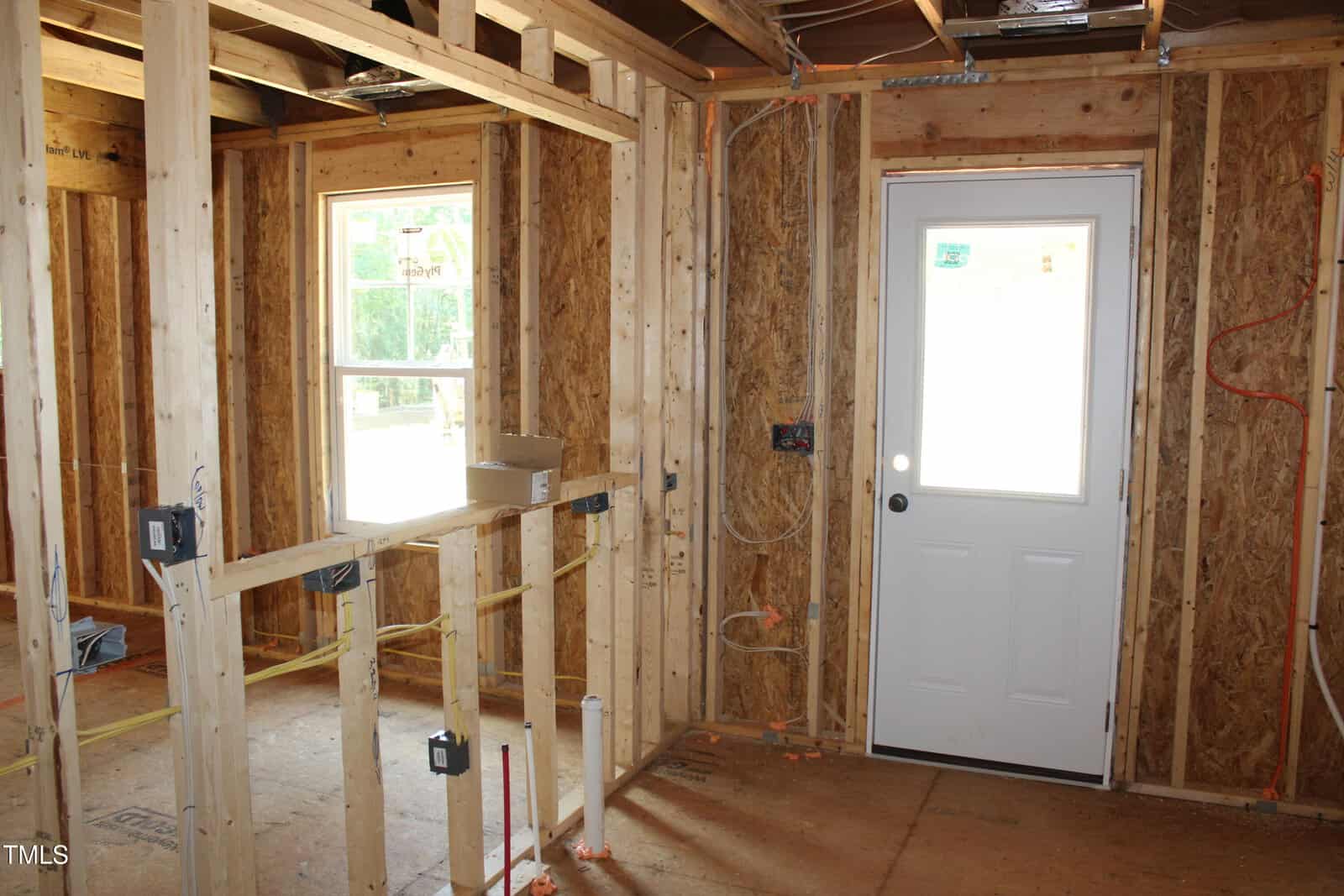 Interior view of a house under construction, showing exposed wooden framing, wiring, and plumbing. A white exterior door and window allow light into the unfinished room. No drywall or insulation is present.