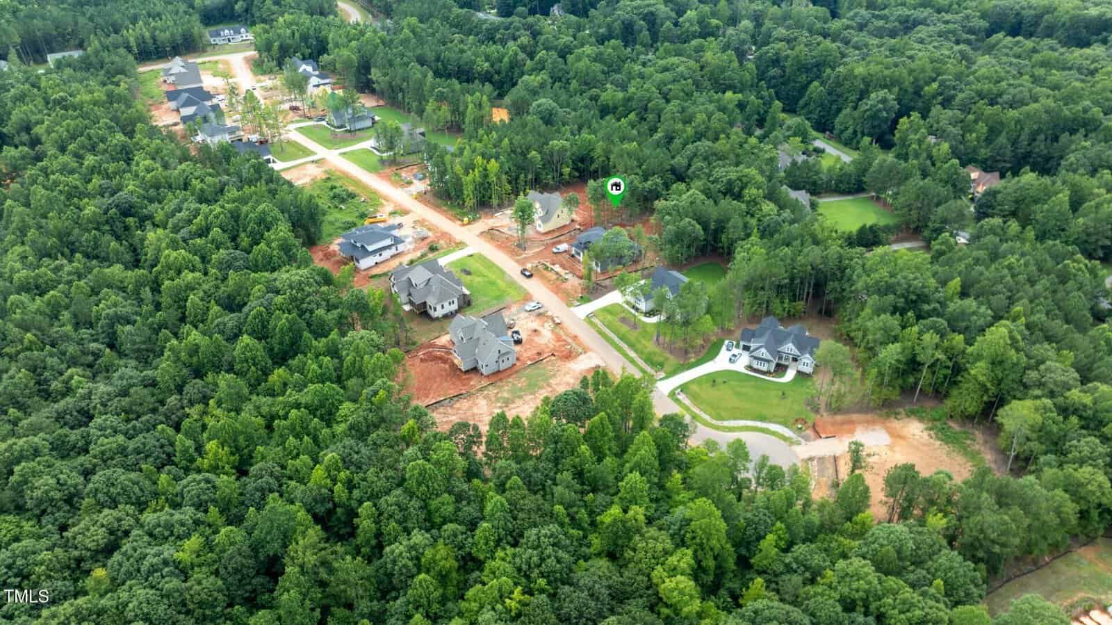 Aerial view of a suburban neighborhood surrounded by dense forest. Several houses are under construction, while some are completed. Roads, cleared lots, and green trees are visible throughout the area.