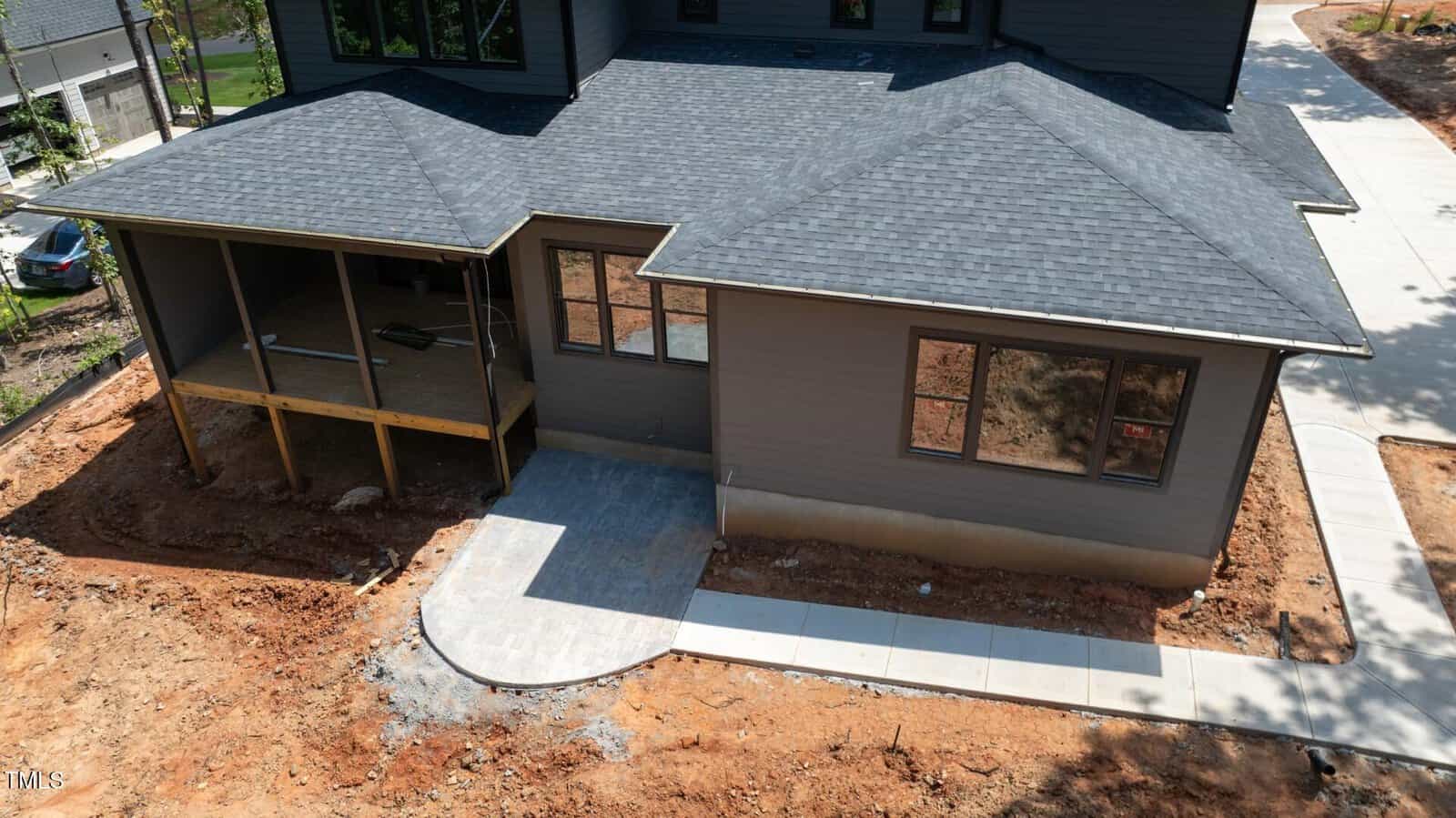 A back view of a modern house under construction showing a covered porch, multiple windows, a paved walkway, and unfinished landscaping with exposed soil.