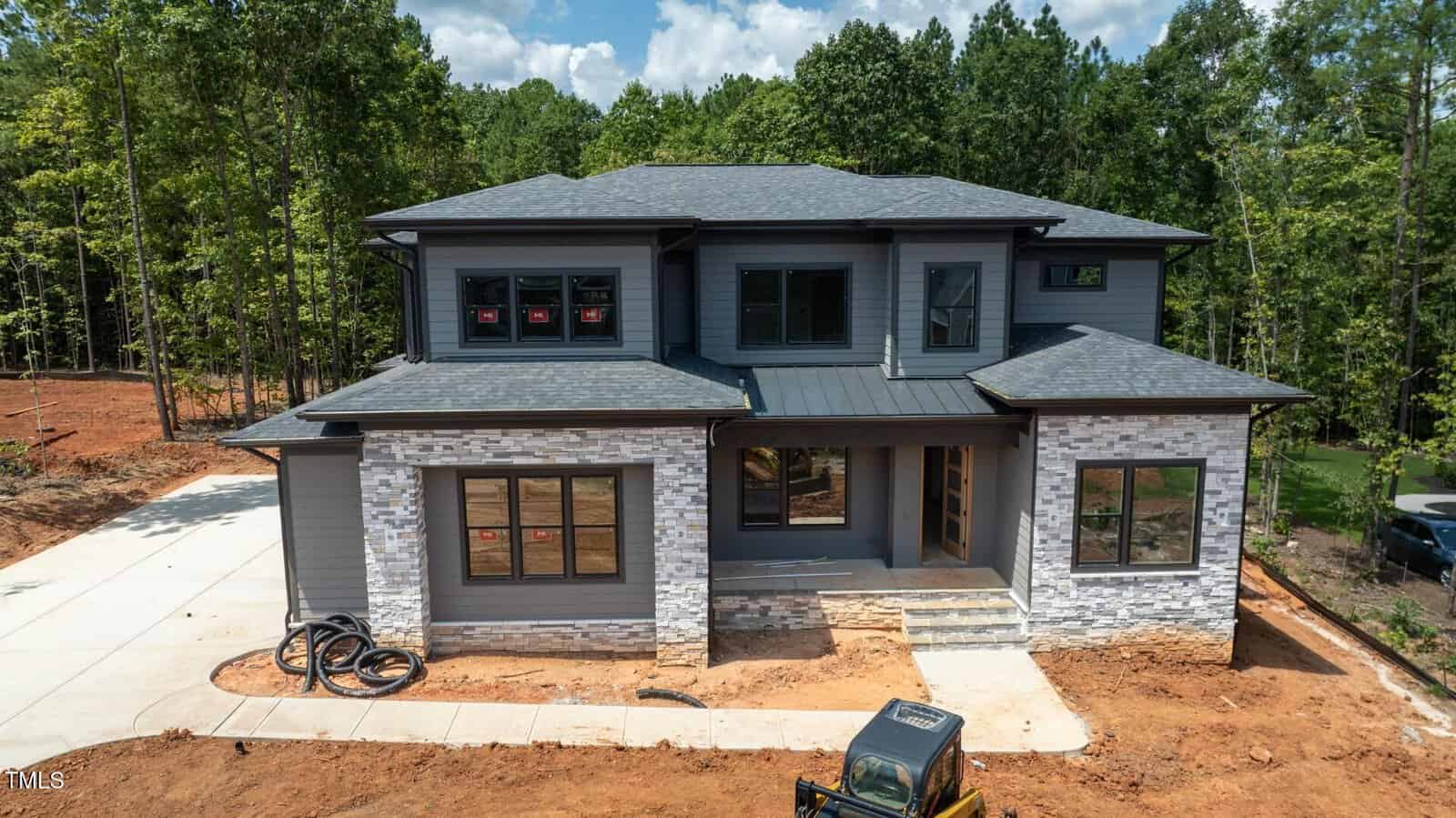 A two-story modern house under construction at 3921 Cedar Knolls Drive in Youngsville features gray siding, stone accents, large windows, a covered front porch, and driveway. Construction materials and equipment are visible on site with trees in the background.
