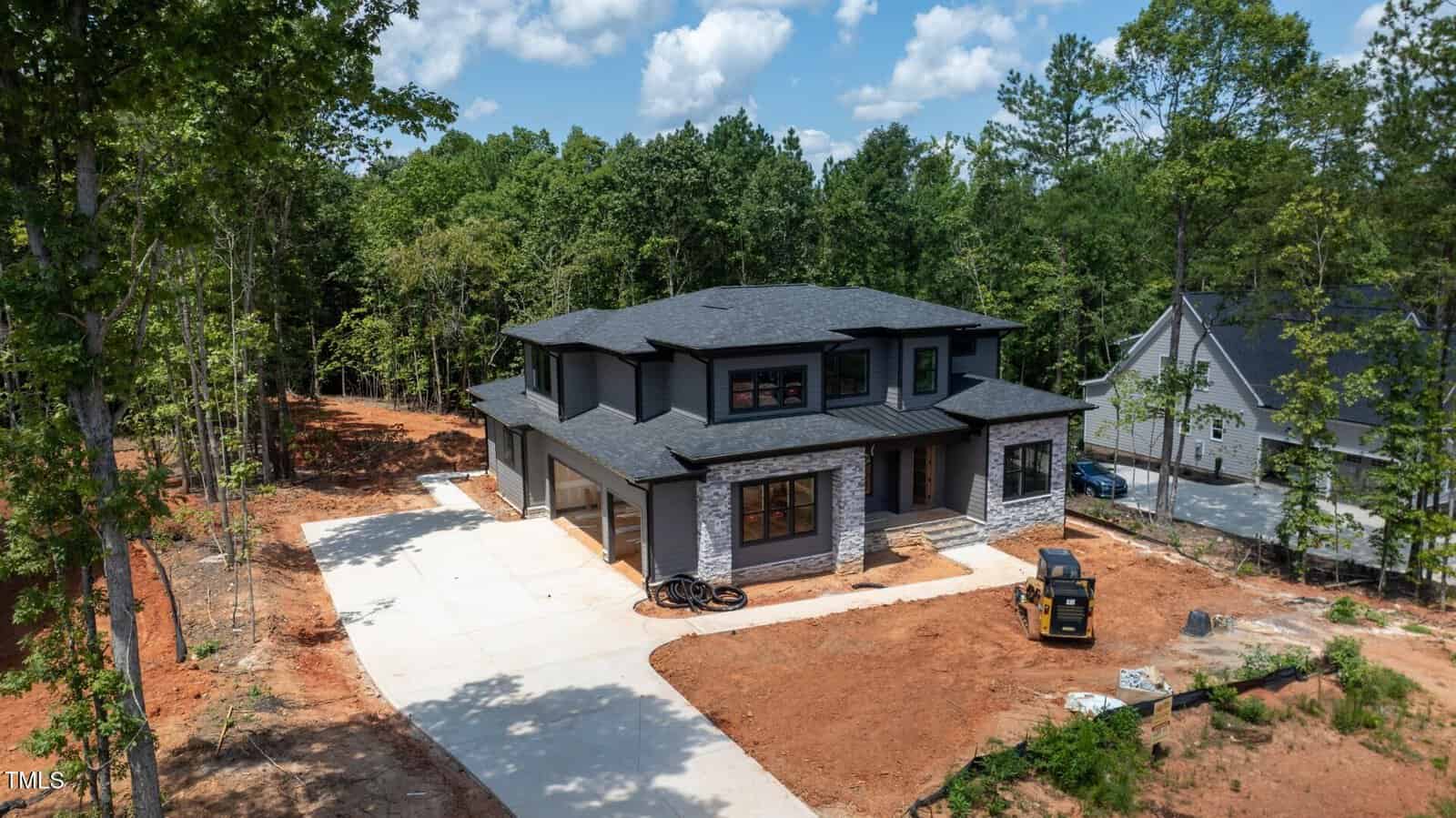 A two-story modern house with gray stone and dark siding sits on a large, newly landscaped lot with bare soil and a paved driveway. Construction materials and a small machine are near the front of the house.