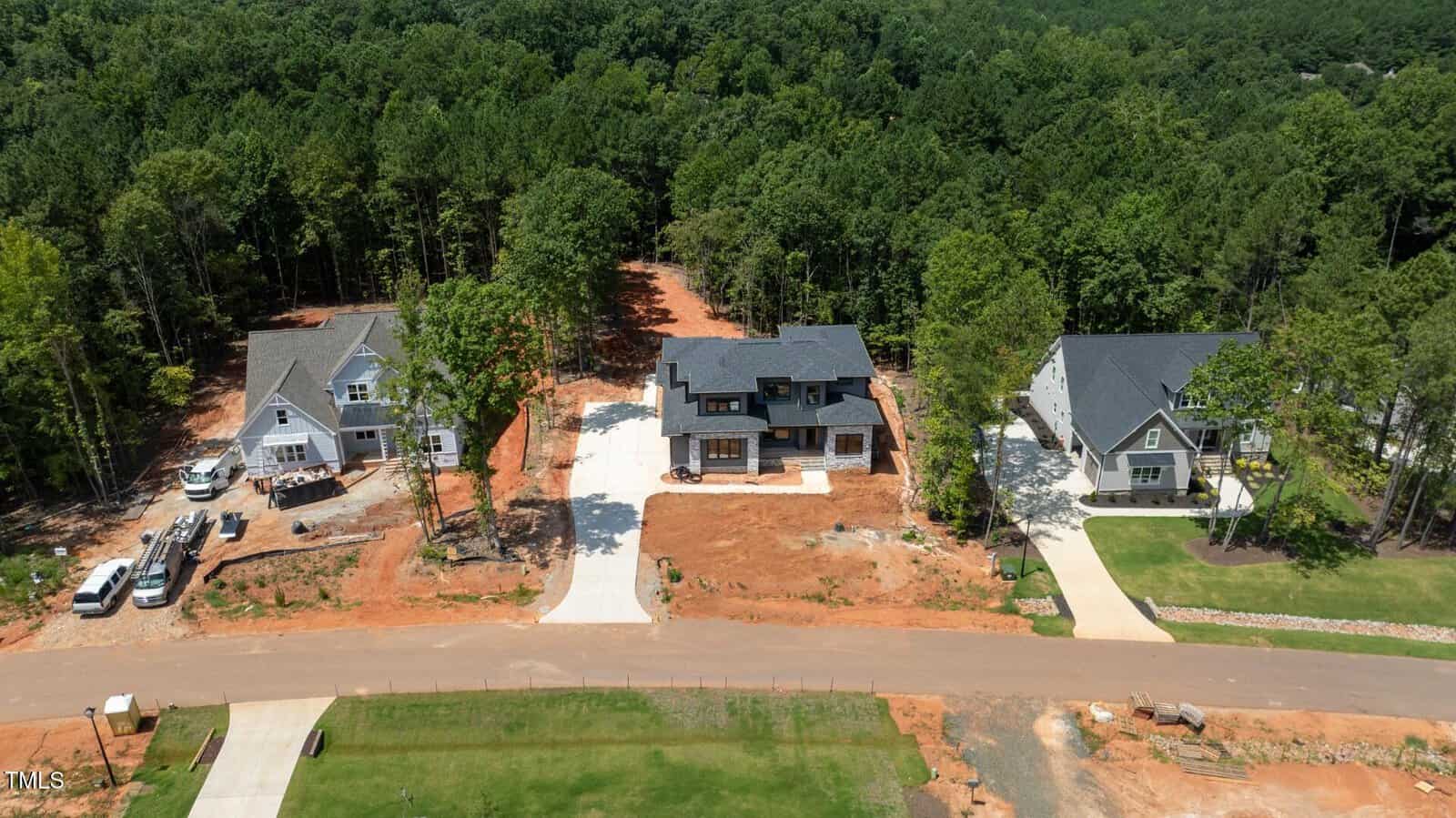 Aerial view of three newly built houses with driveways on a dirt lot, surrounded by trees and bordered by a paved road in a suburban neighborhood.