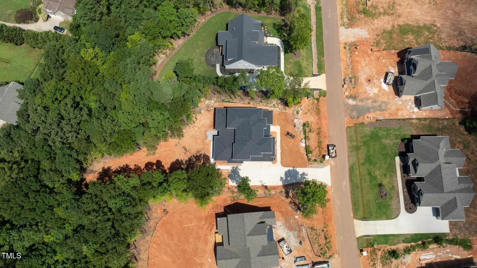Aerial view of a suburban neighborhood under development, showing several completed houses, driveways, green lawns, and surrounding trees, with areas of exposed red soil where construction is ongoing.