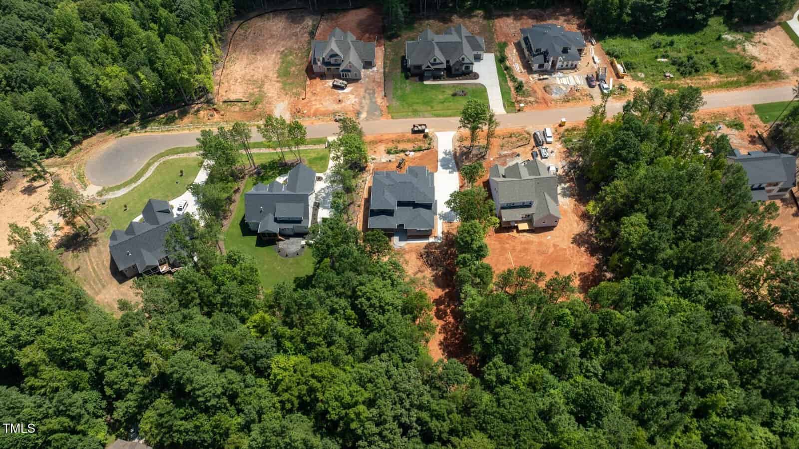 Aerial view of several houses in a residential neighborhood with some homes under construction, surrounded by trees and patches of red soil. A road runs horizontally through the center of the image.