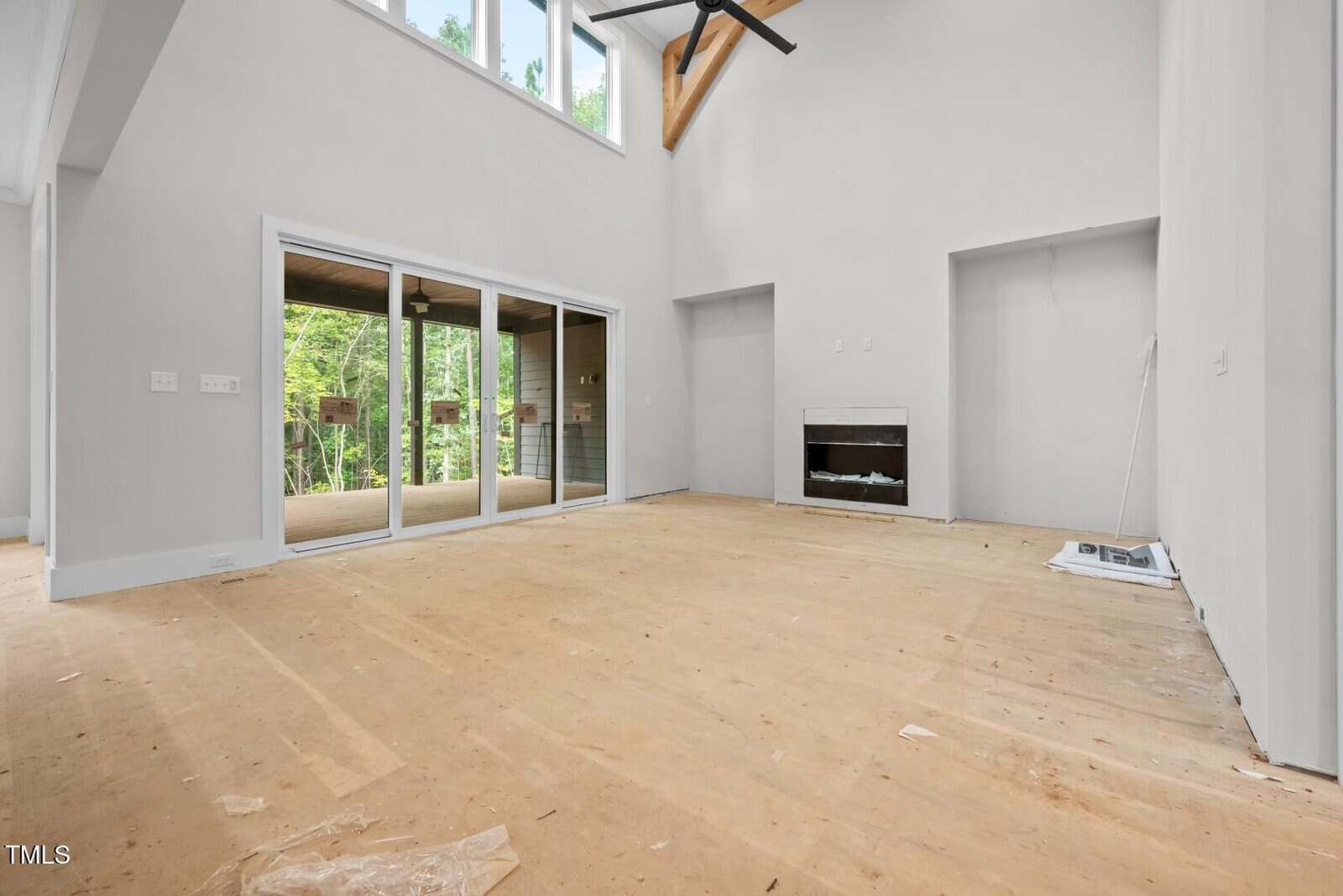 A large, empty living room with unfinished floors, white walls, a built-in fireplace, tall ceiling, ceiling fan, and glass sliding doors leading to a covered outdoor area. Construction materials are on the floor.