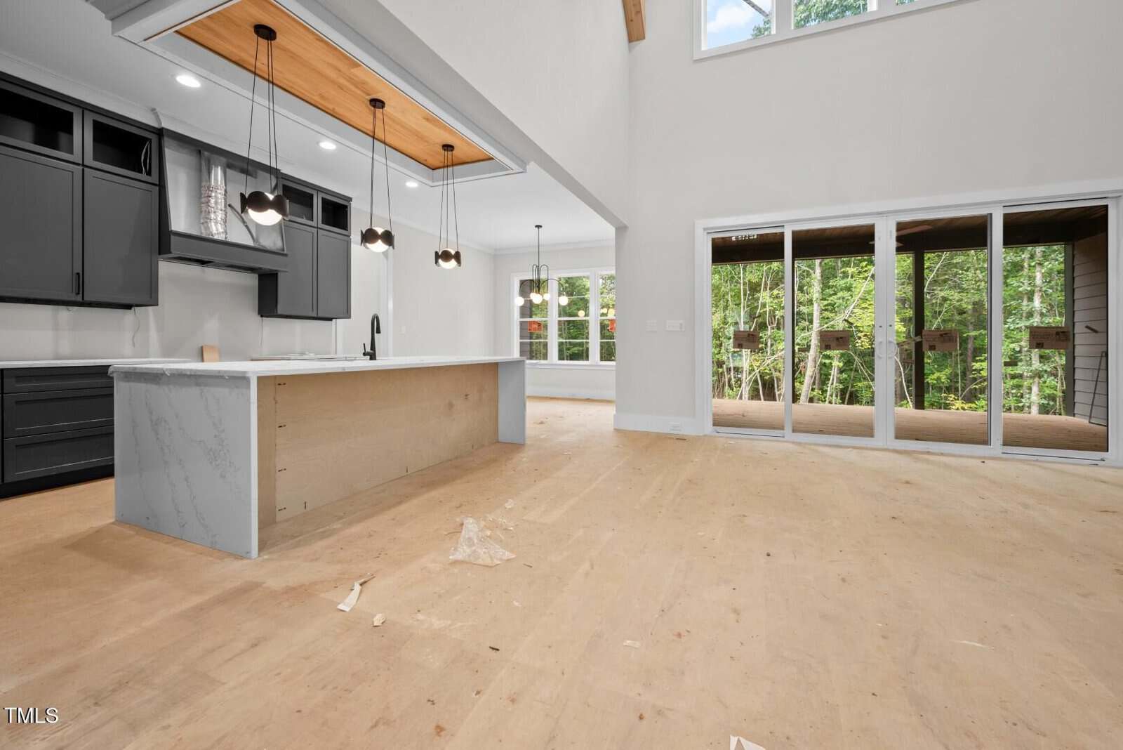 Unfurnished kitchen and living area with light wood floors, dark cabinets, a large kitchen island, pendant lights, and sliding glass doors leading to a screened porch overlooking trees. Construction debris is on the floor.